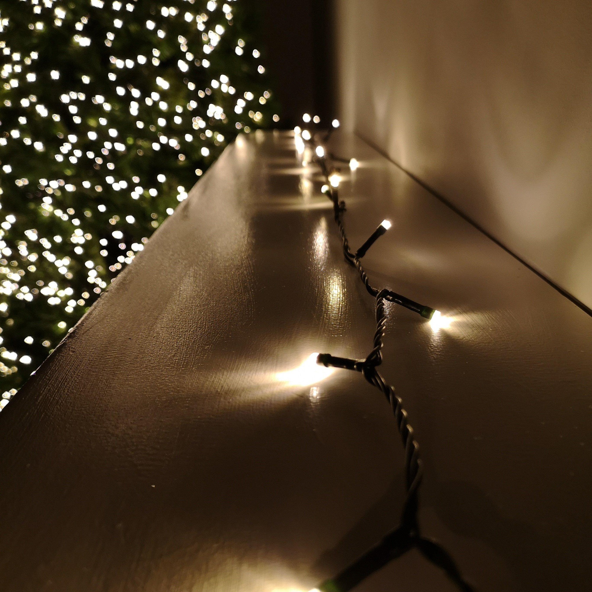 Warm white LED string lights with a black wire, featuring 720 bulbs, designed for Christmas decoration, with a timer function, illuminating a blurred green tree in the background.