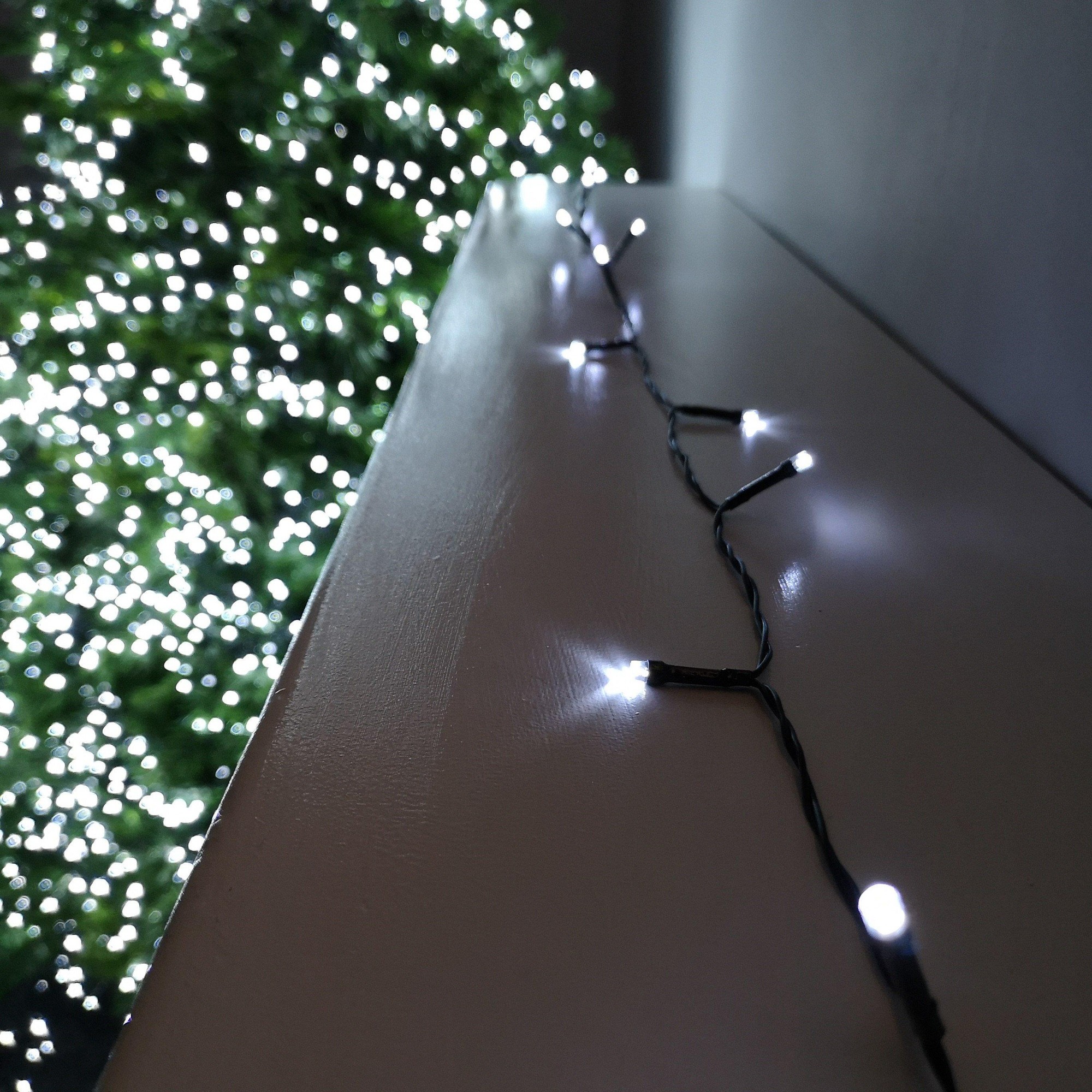 Battery-operated 100m string lights featuring 100 cool white LED bulbs on a black wire, elegantly draped along a surface with a blurred green tree in the background.
