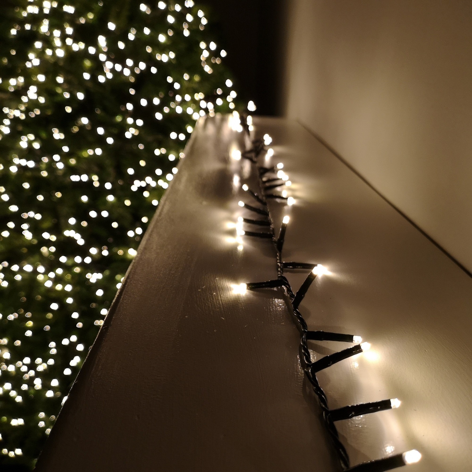 Warm white LED string lights with a black wire, featuring multiple action settings and a timer, displayed along a ledge with a softly lit Christmas tree in the background.