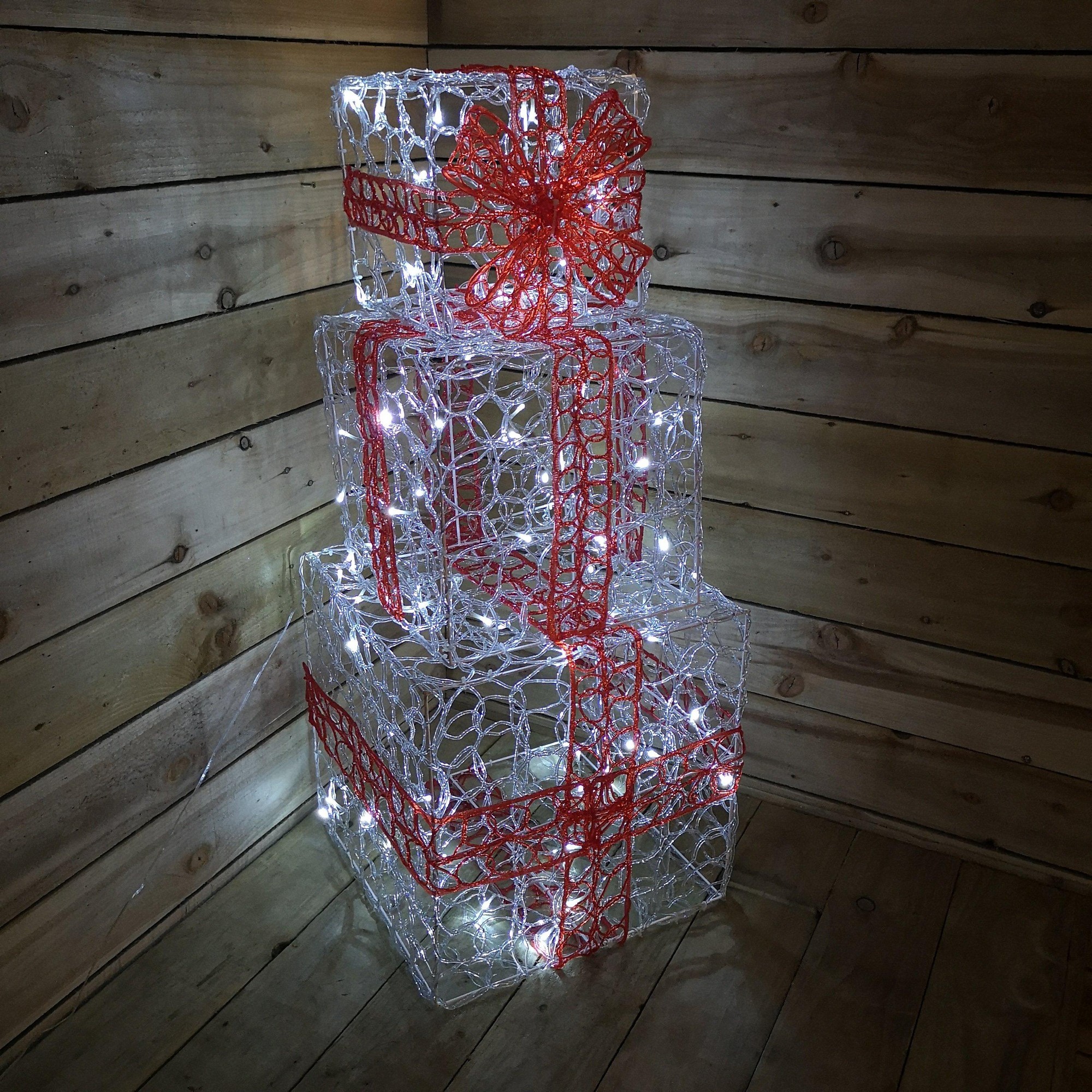 Three stacked acrylic gift parcels in white, adorned with red bows, illuminated by warm white LED lights, set against a wooden backdrop.