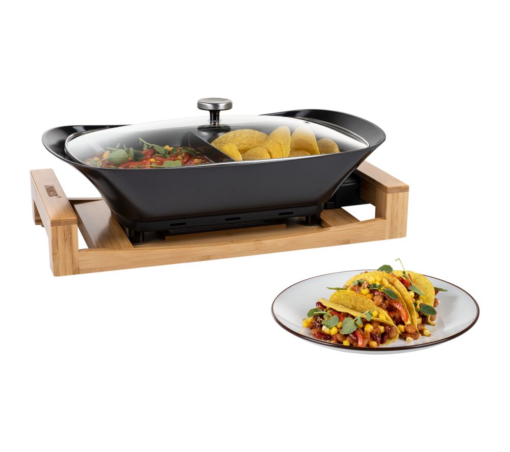 Black multi-cook tabletop appliance with a glass lid, wooden base, and a side of tacos on a white plate, featuring colorful vegetables inside the cooking pan.