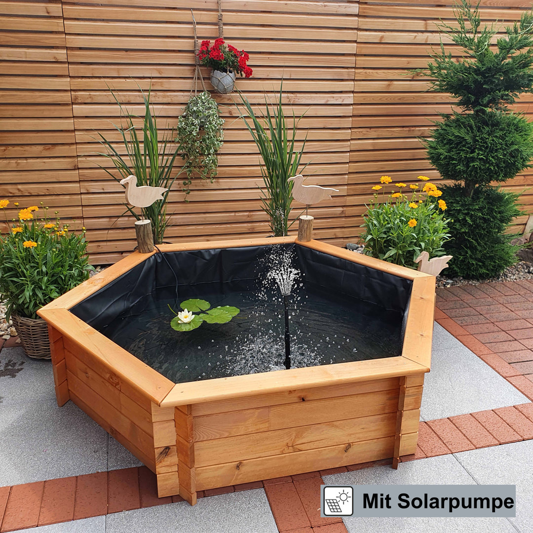 Brown hexagon-shaped garden pond set with a black liner, featuring a solar fountain, decorative ducks, and surrounded by vibrant flowers and greenery.