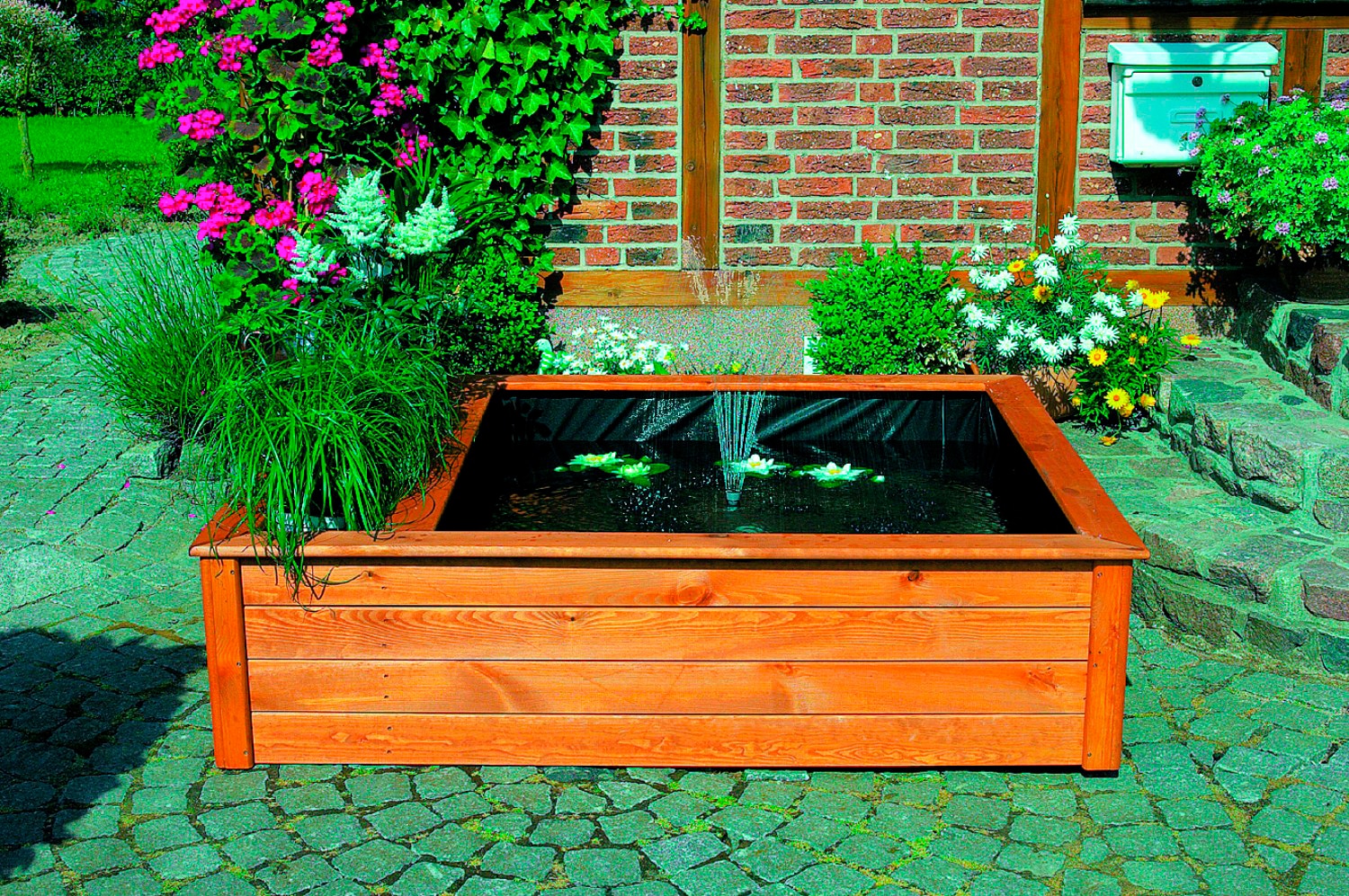Raised square garden pond kit in brown wood, featuring a black liner, water fountain, and planting zone, surrounded by vibrant flowers and greenery.