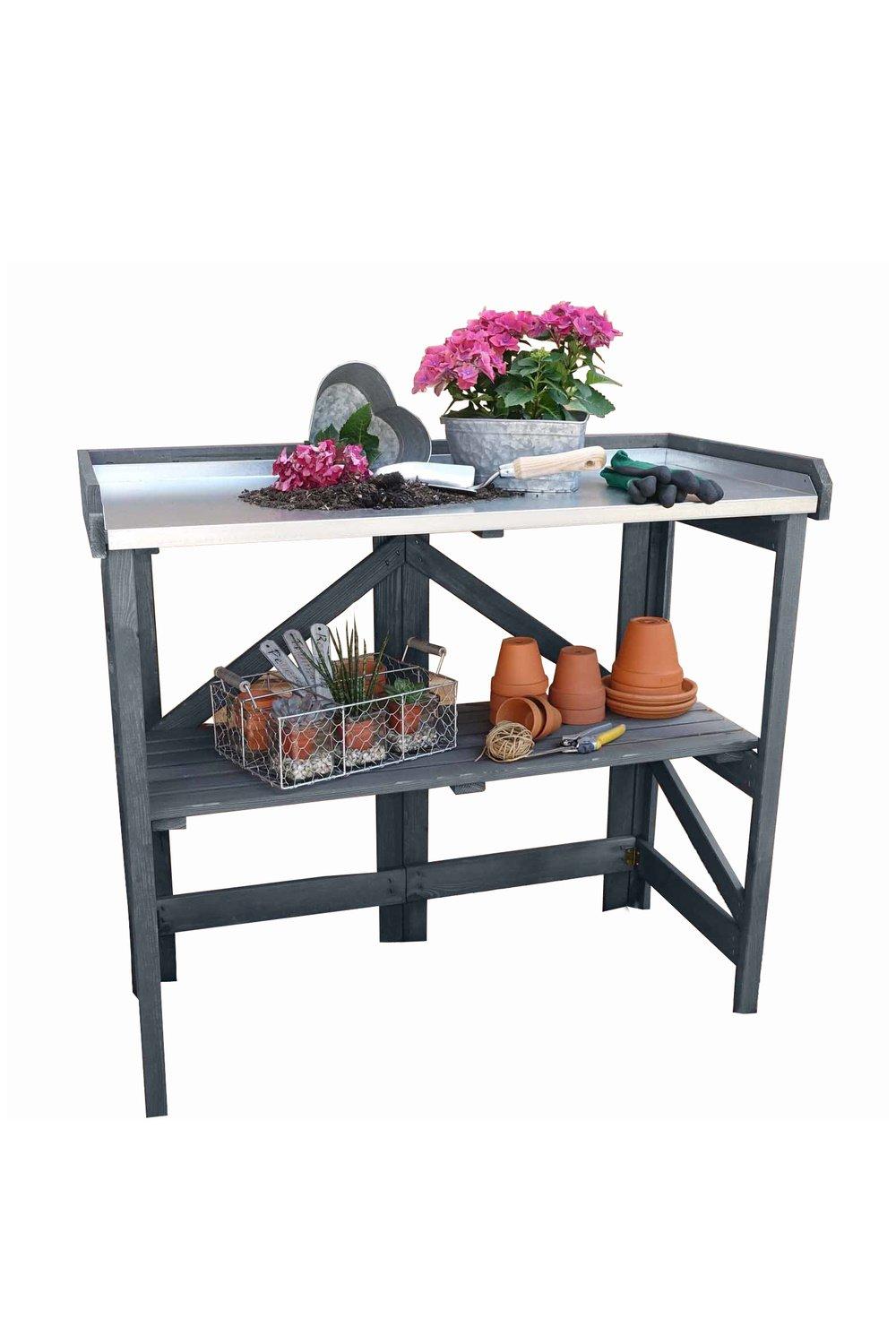 Grey wooden garden potting table featuring a zinc-plated worktop, lower shelf with terracotta pots, gardening tools, and a planter with pink flowers.