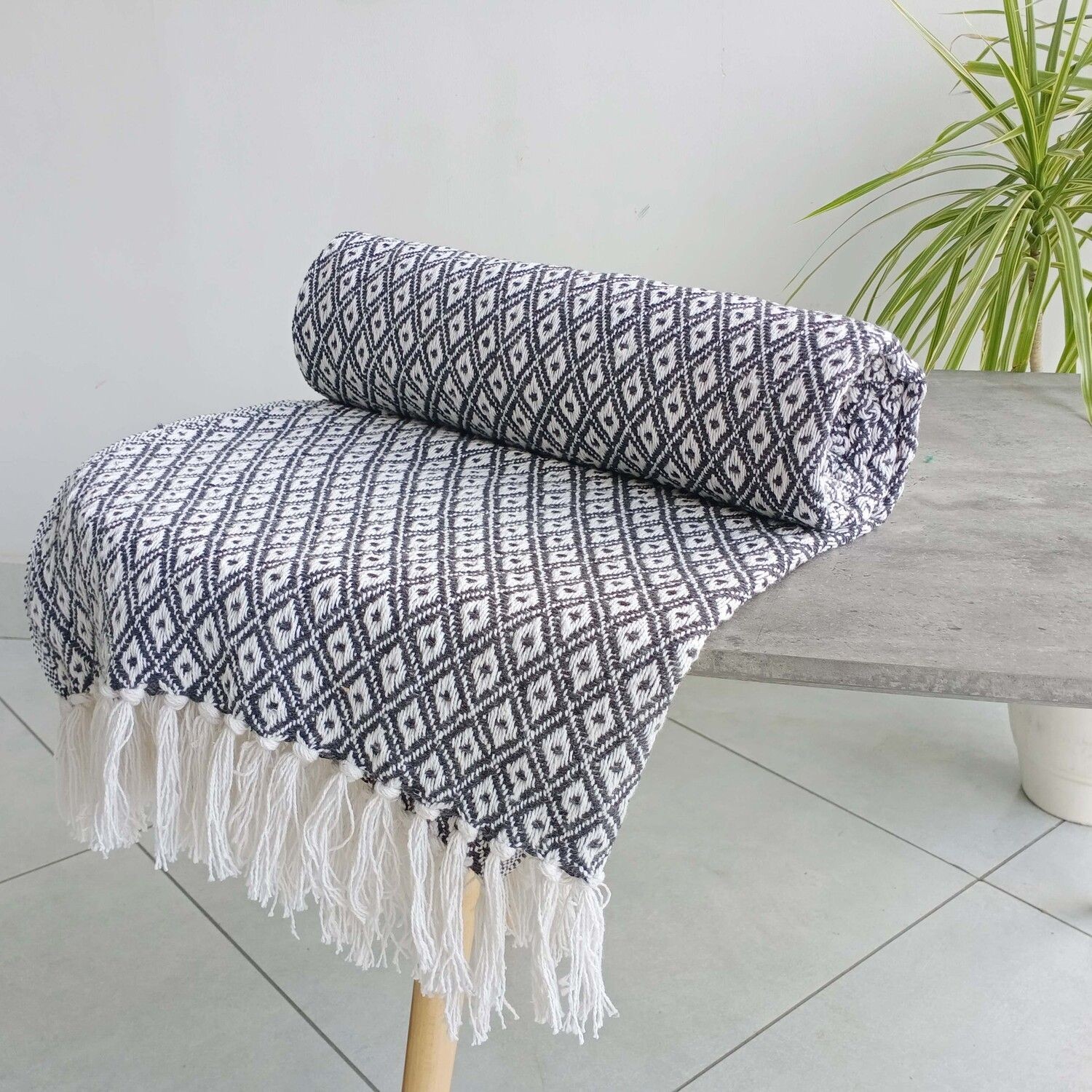 Charcoal and grey cotton throw featuring a geometric diamond pattern, rolled neatly with fringed edges, displayed on a wooden stool against a light background.
