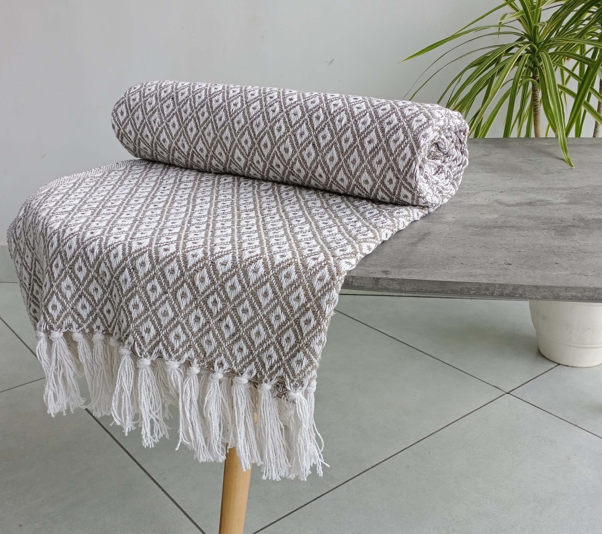 Natural and brown cotton throw featuring a geometric diamond pattern, rolled neatly with fringed edges, placed on a modern table beside a potted plant.