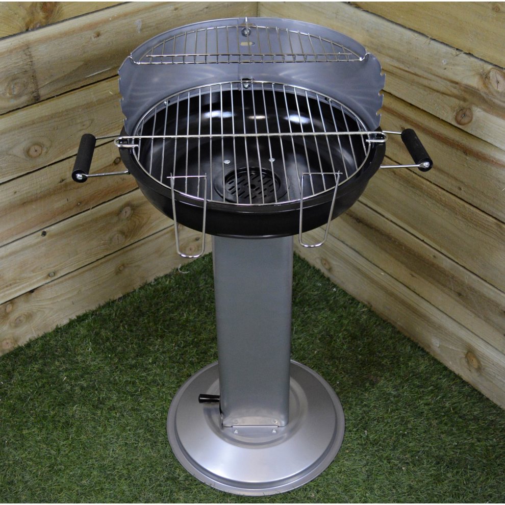 Half round black barbecue on a silver stand with a vented design, featuring a chrome grill and wooden handles, set against a wooden backdrop and grass-like surface.