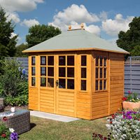 A wooden summerhouse with a light brown finish, featuring large glass windows, a sloped green roof, and double doors, surrounded by colorful flowers and greenery.