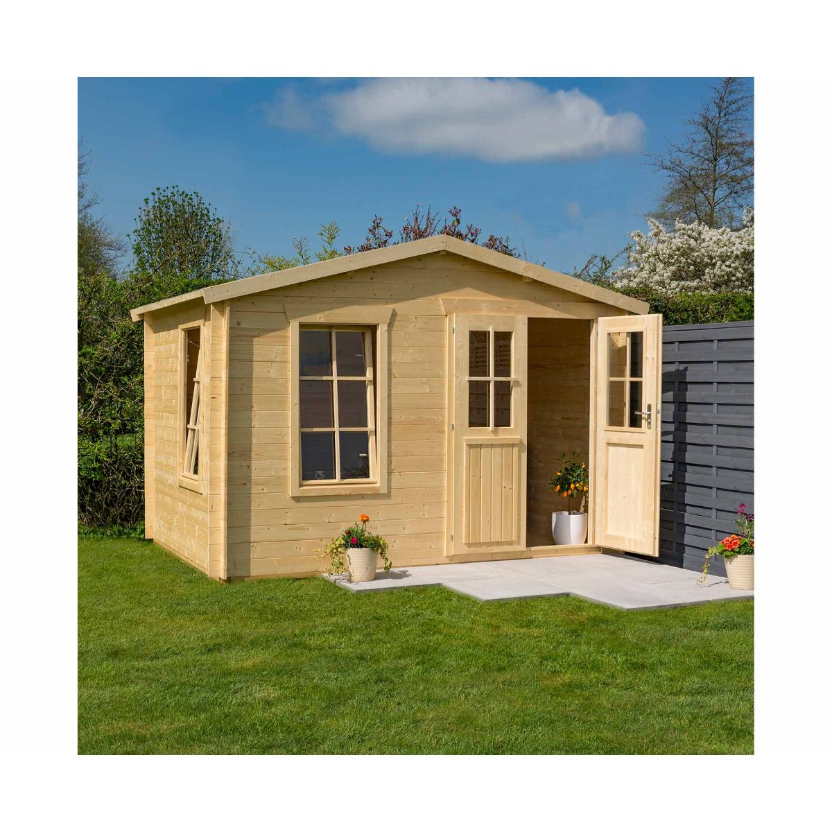 Light wooden log cabin garden studio with a sloping roof, double doors, two windows, and flower pots on a stone patio, surrounded by green grass and trees.