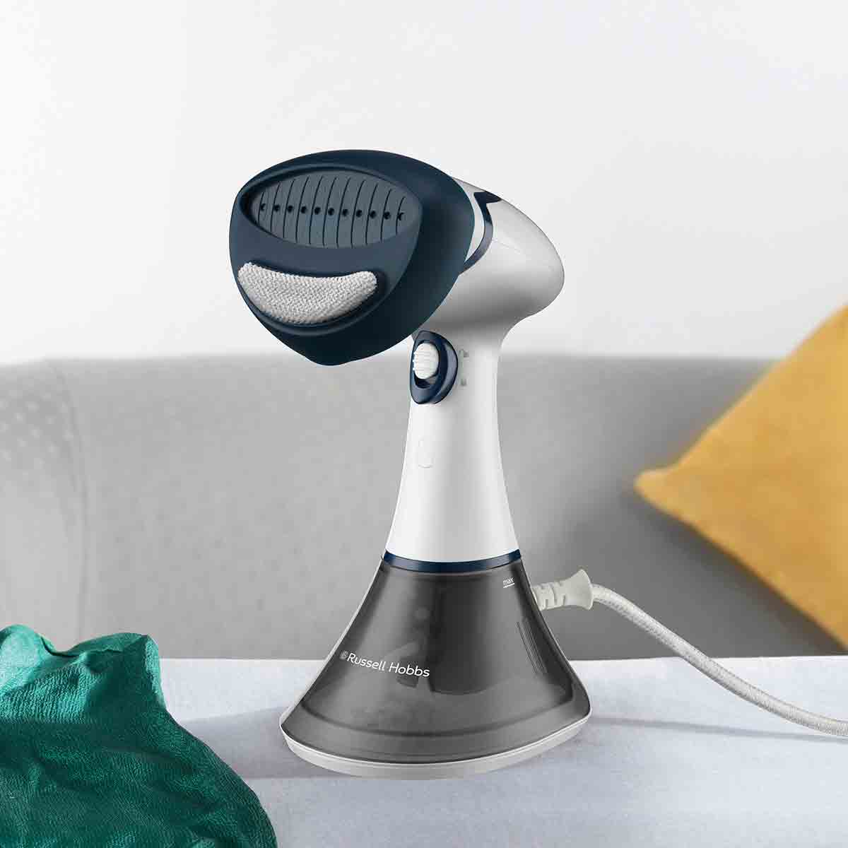 White and navy handheld garment steamer with a textured steam head, a transparent water tank, and a curved base, resting on a light-colored fabric surface beside a green cloth and yellow cushion.