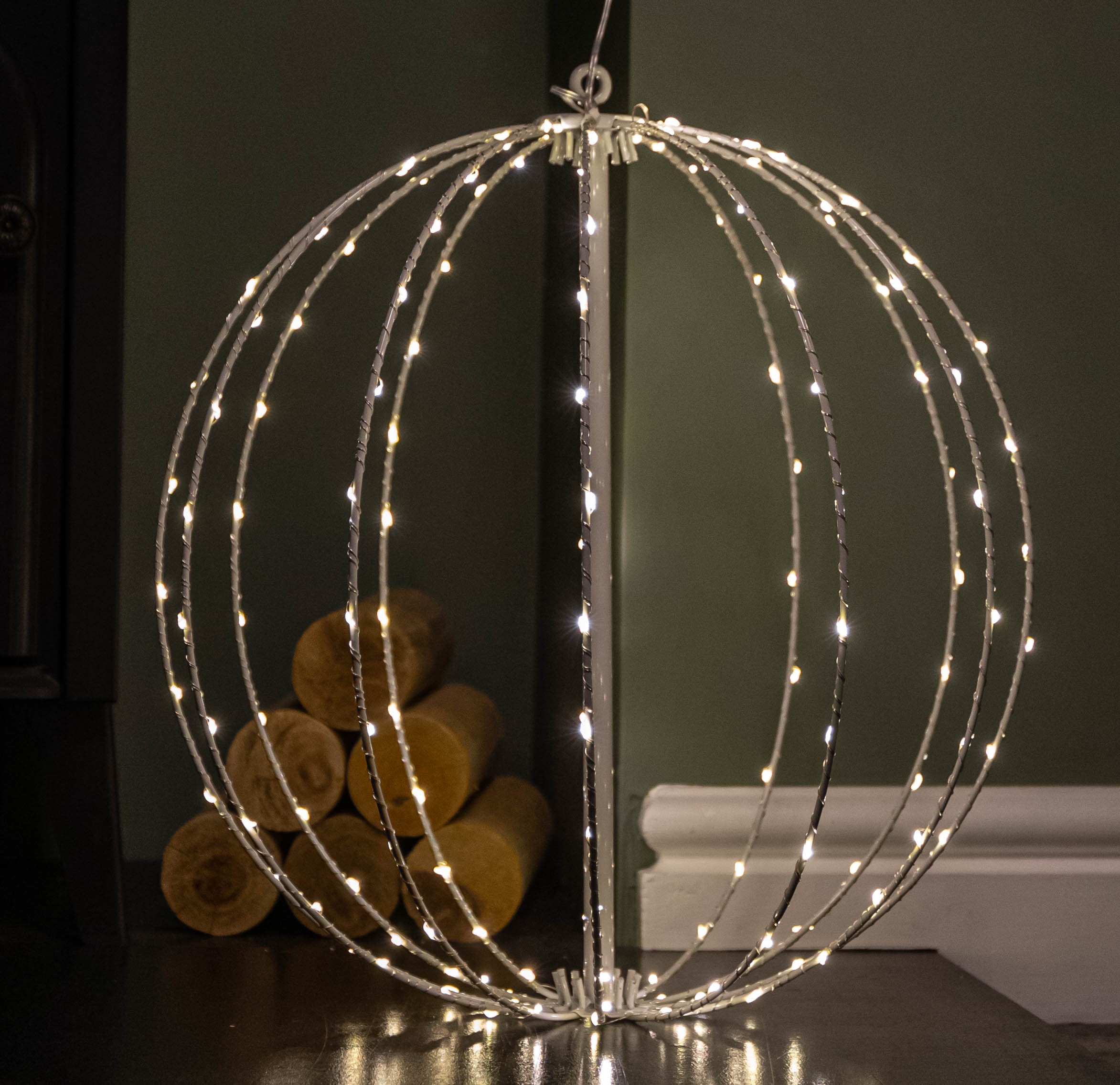 30cm white metal folding Christmas ball featuring 108 warm white LEDs, with a spherical design and illuminated strands, set against a dark green wall and wooden logs.