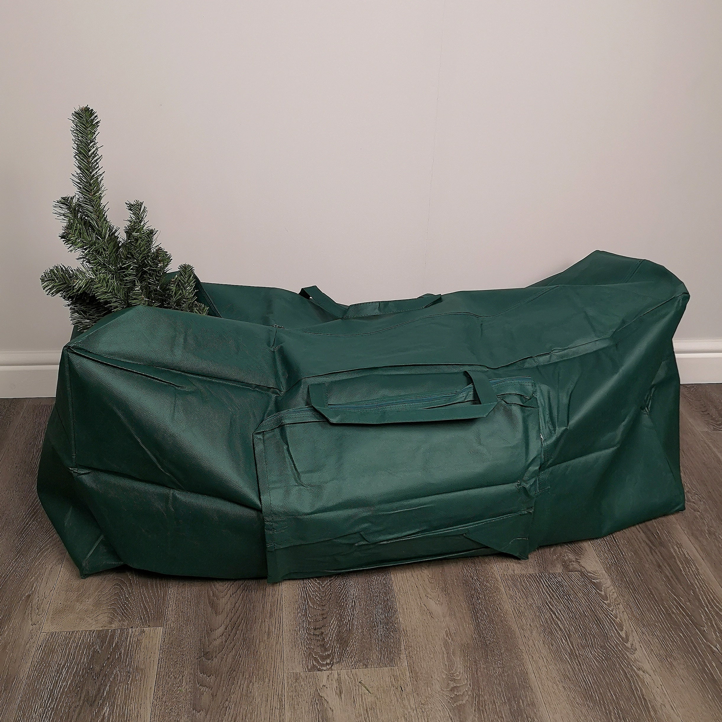 Green storage bag for a Christmas tree, made of durable material, featuring a zip closure and a carry handle, with a small section of tree visible at one end.
