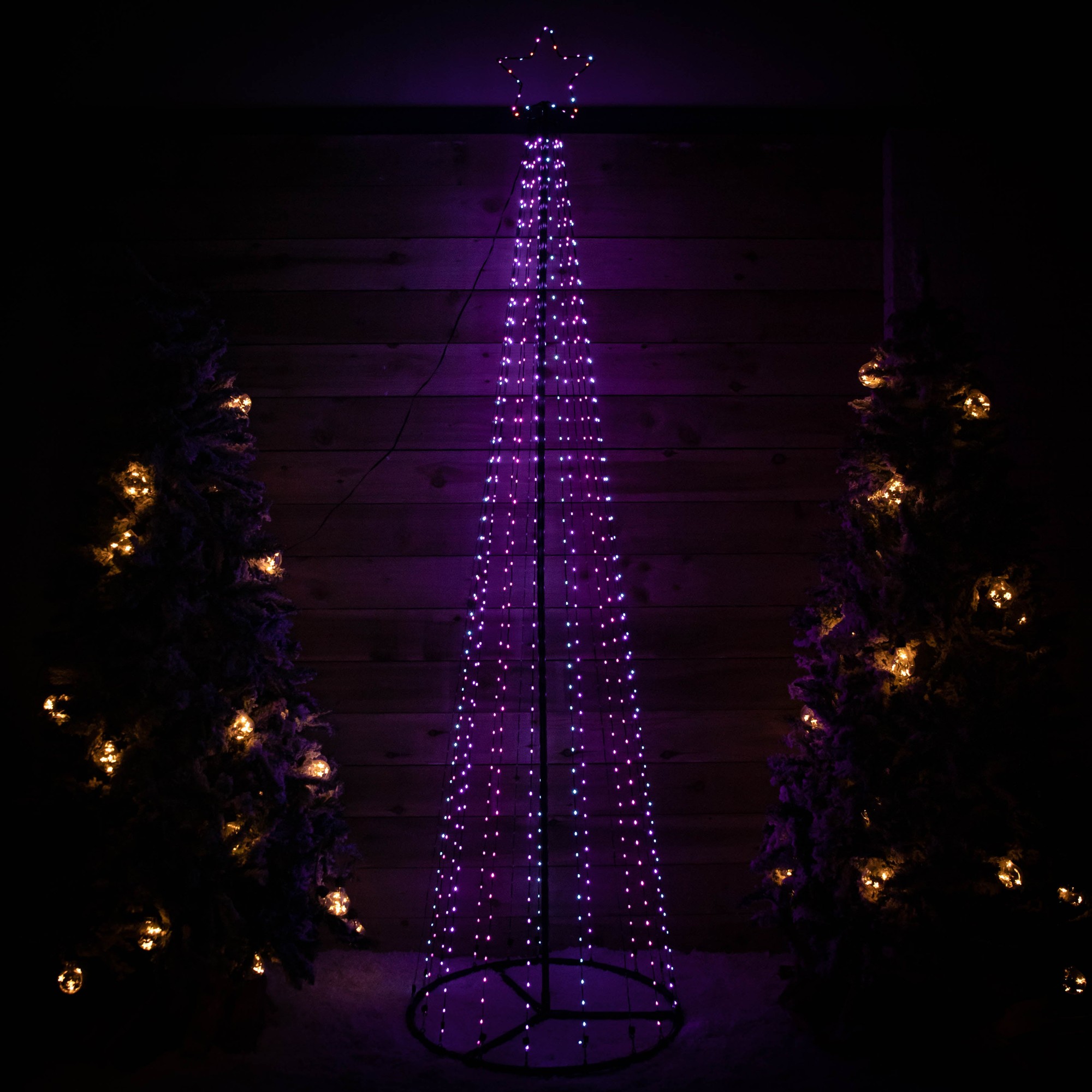 A tall, cone-shaped Christmas tree made of metal with 1374 multi-action rainbow LEDs, illuminated in purple, featuring a star at the top and surrounded by decorated trees.