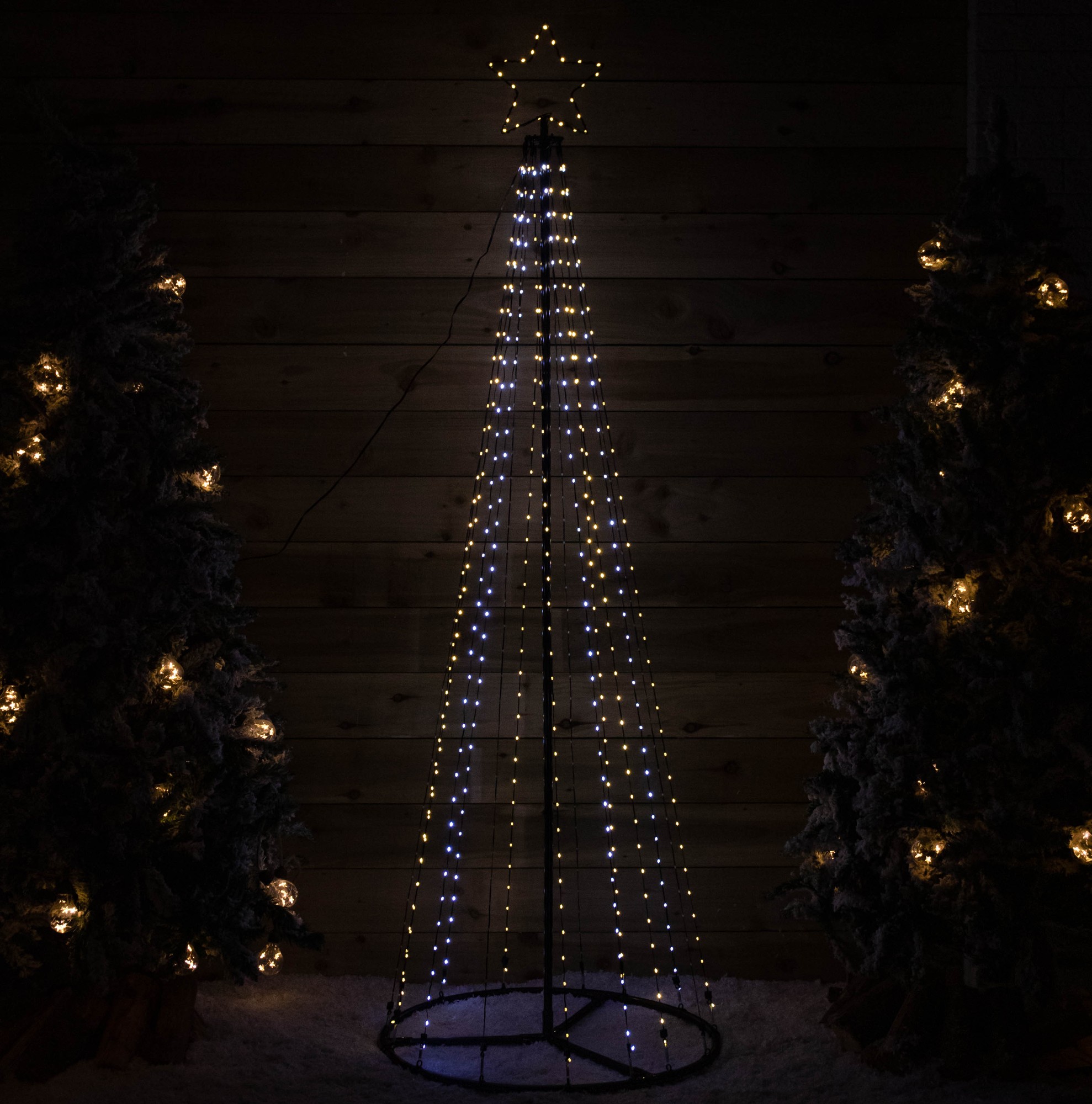A tall, cone-shaped Christmas tree made of metal, adorned with 1374 multi-action white and warm white LEDs, topped with a star, set against a wooden backdrop and surrounded by lit greenery.