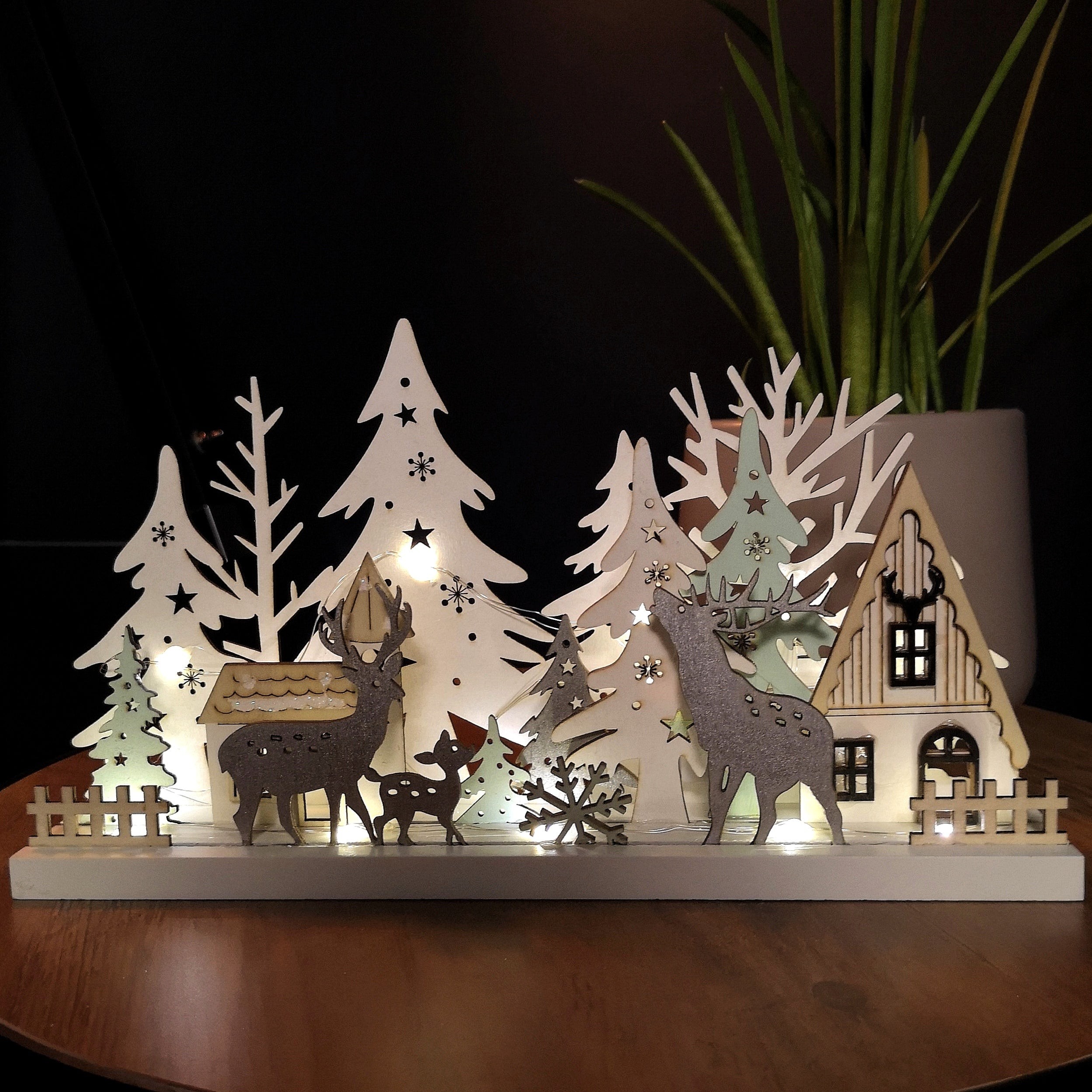 30cm wooden Christmas village scene featuring white trees, reindeer, and a house, illuminated by warm white LEDs, creating a festive ambiance.