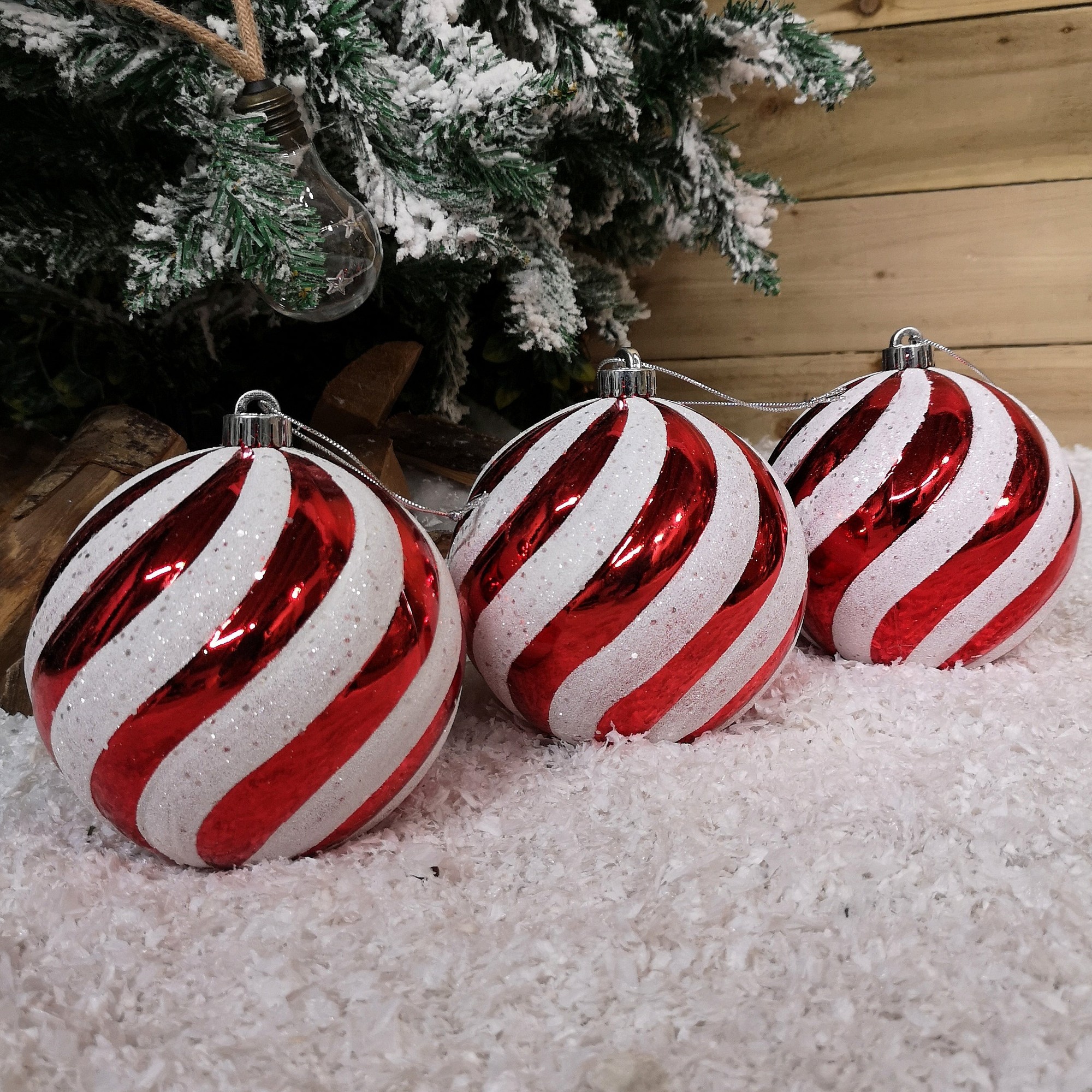 Three large shatterproof Christmas baubles in red and white, featuring a spiral design with glitter accents, resting on a snowy surface near a decorated tree.