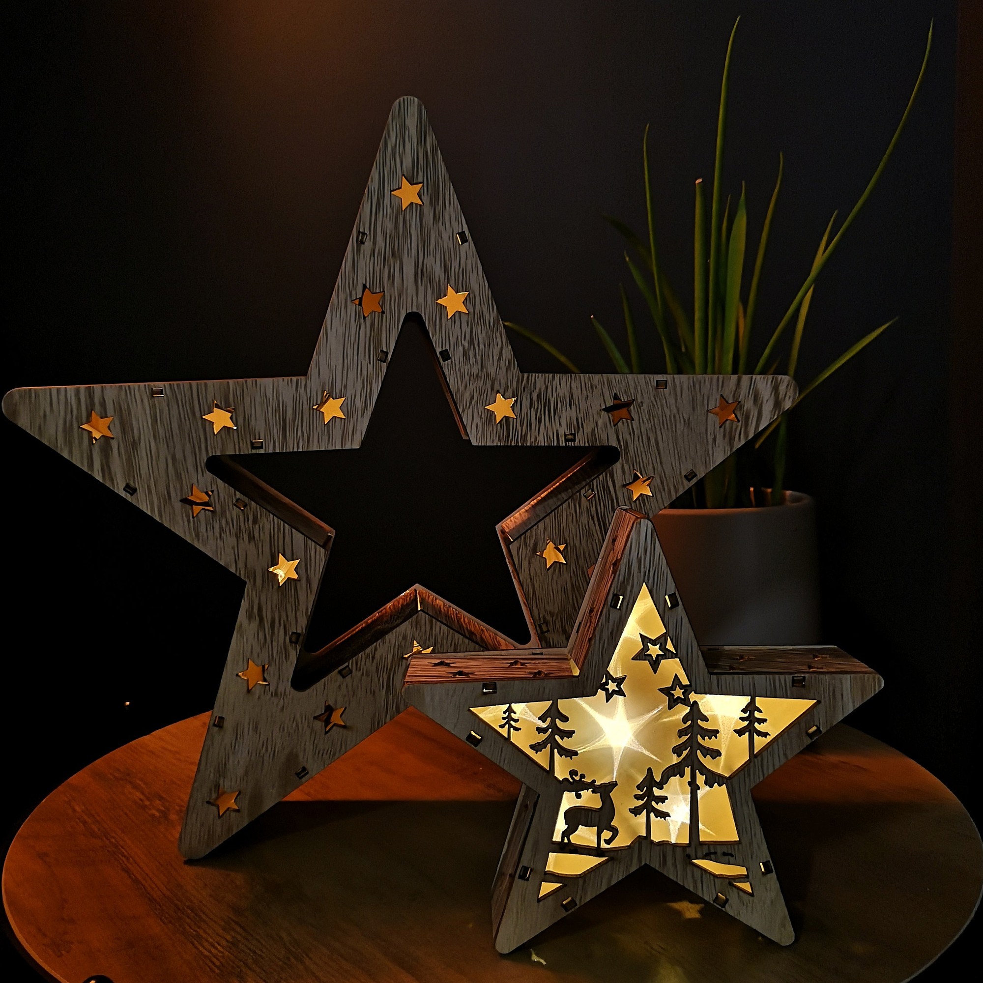 Set of 2 wooden Christmas stars in rustic brown, featuring cut-out designs of trees and deer, illuminated by warm white LEDs, displayed on a wooden surface.