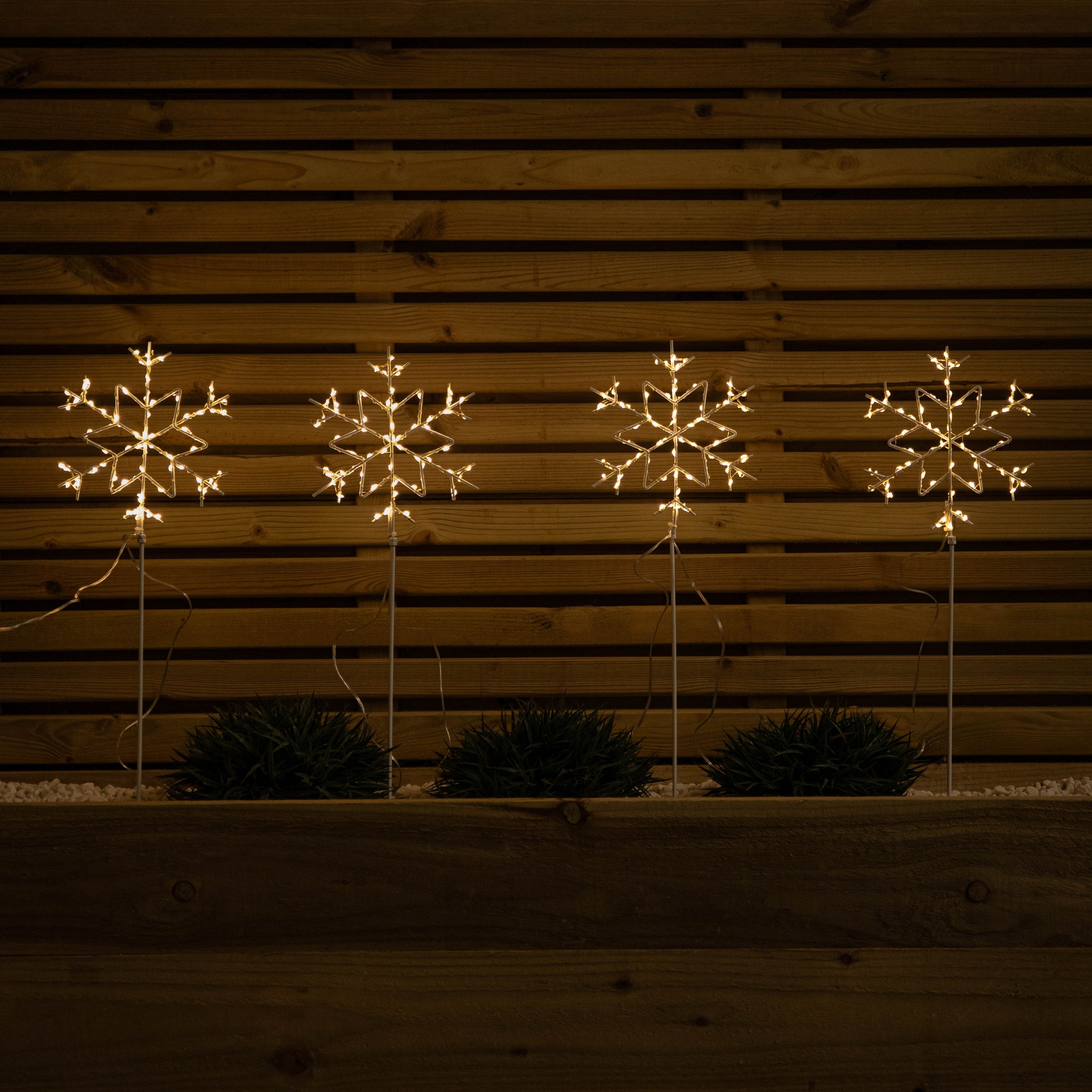 Set of 4 white snowflake path lights, each 60cm tall, adorned with 260 warm white LEDs, mounted on slender stakes, displayed against a wooden backdrop.