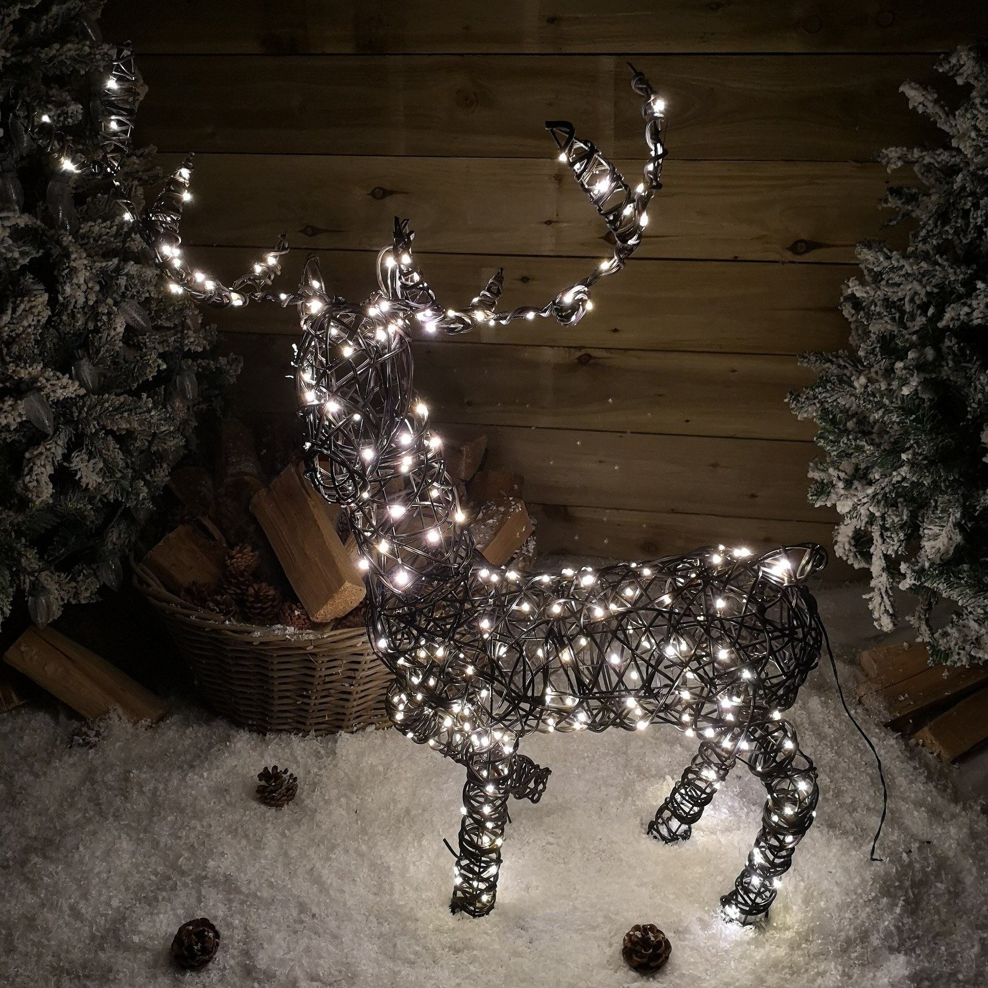 99cm tall wicker reindeer Christmas decoration, wrapped in warm white LED lights, set against a snowy backdrop with pinecones and frosted greenery.