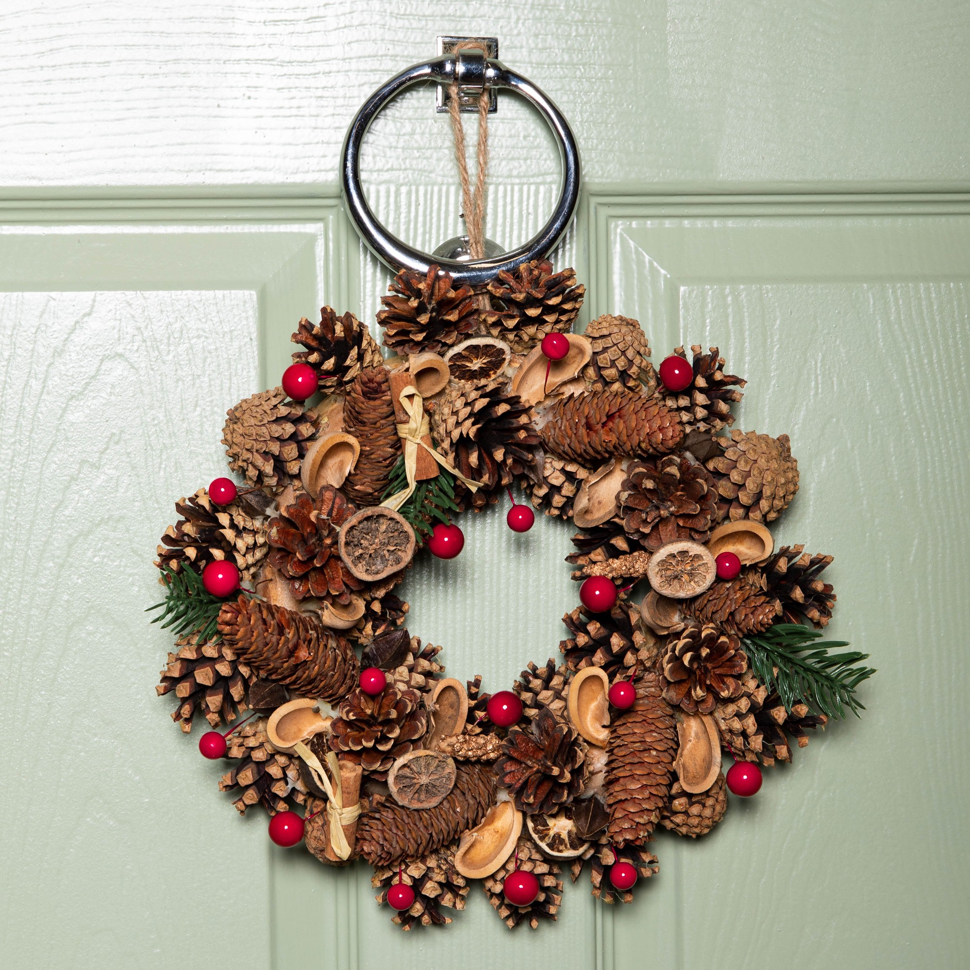Winter spice wreath featuring pine cones, red berries, and dried fruit, adorned with a metal ring for hanging, set against a green door.