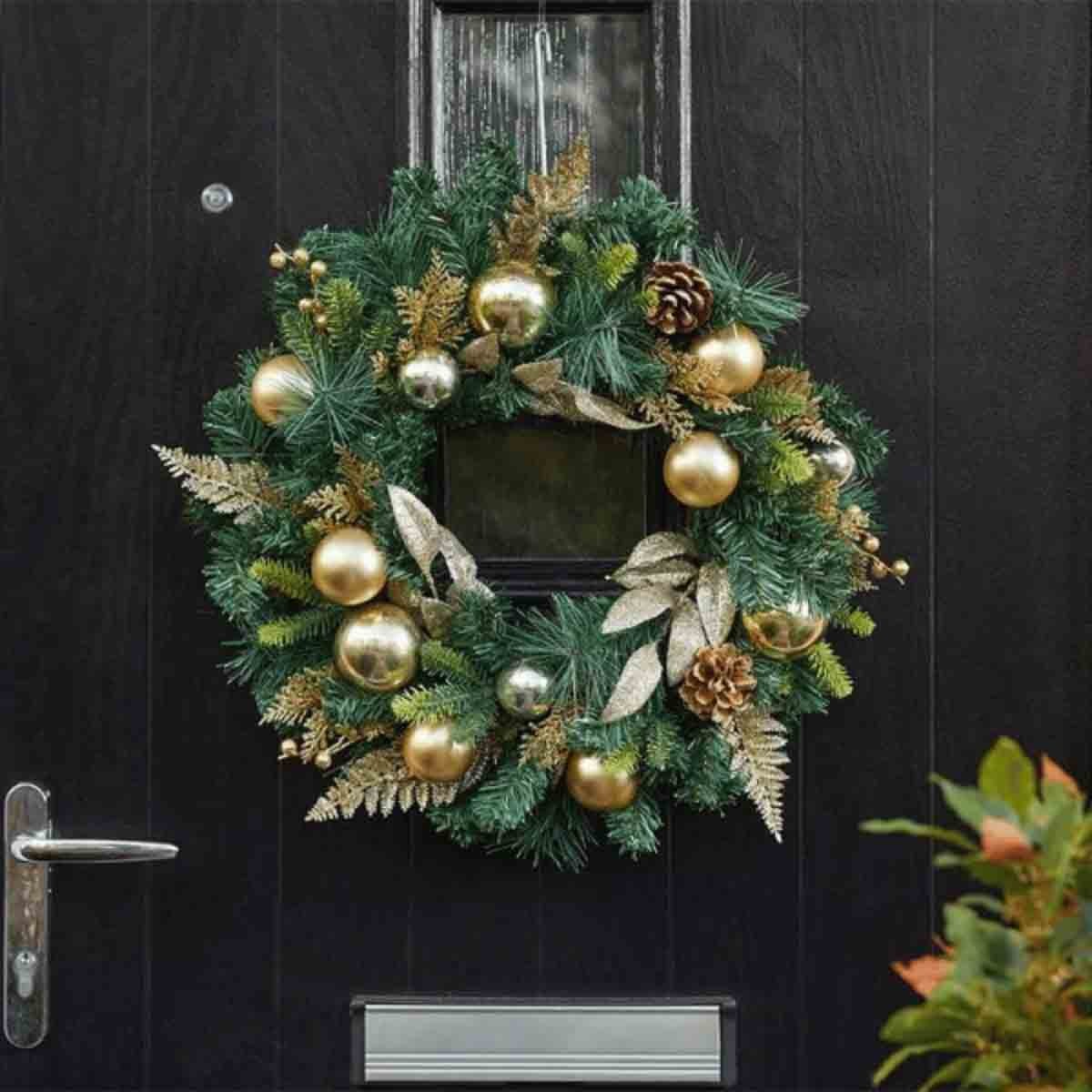A 40cm gold Christmas wreath featuring green pine branches, shiny gold baubles, pinecones, and decorative leaves, hanging on a dark door with a small window.