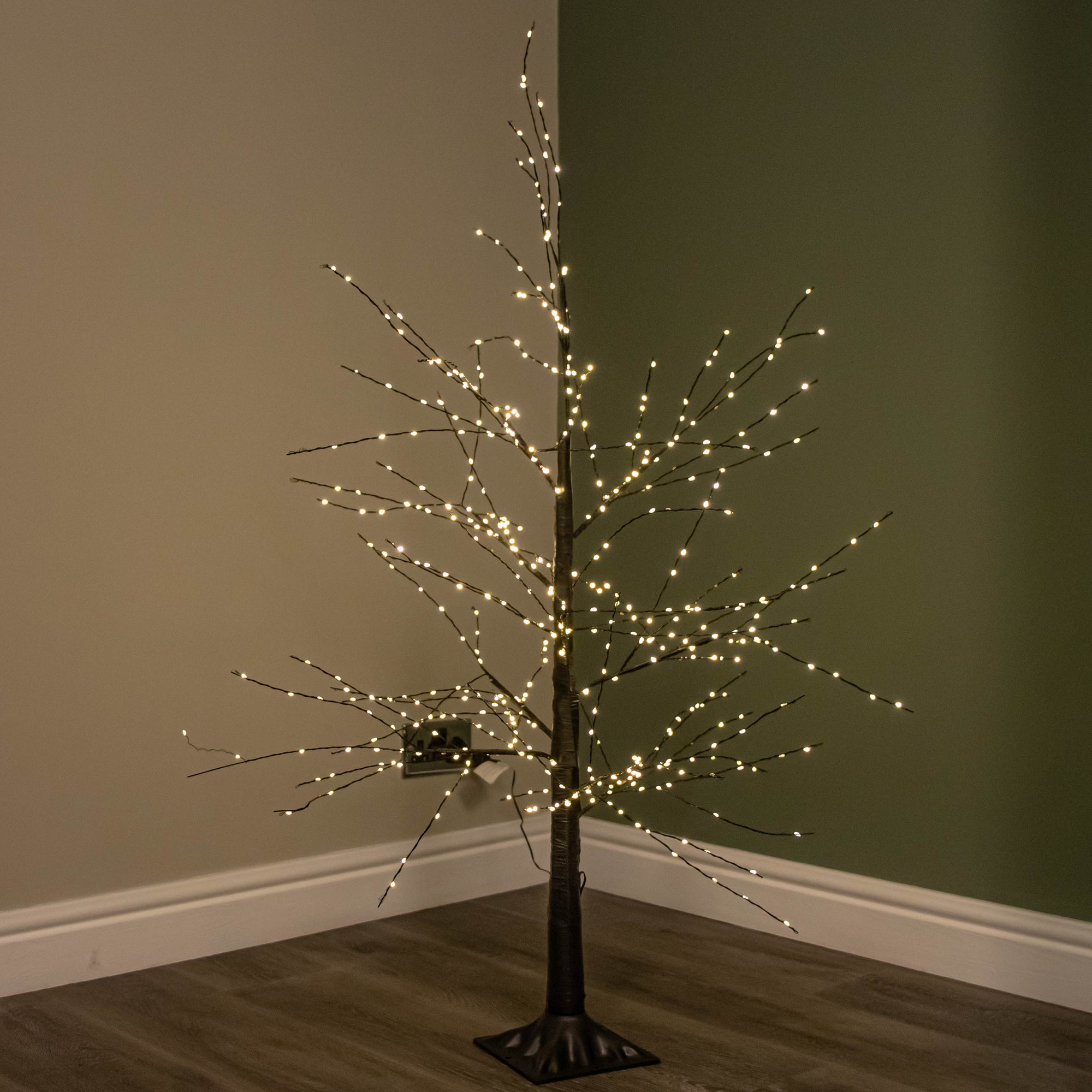 4ft black micro dot Christmas tree with a slender trunk and bare branches, adorned with 500 warm white LEDs that twinkle and shine, set against a muted green wall.