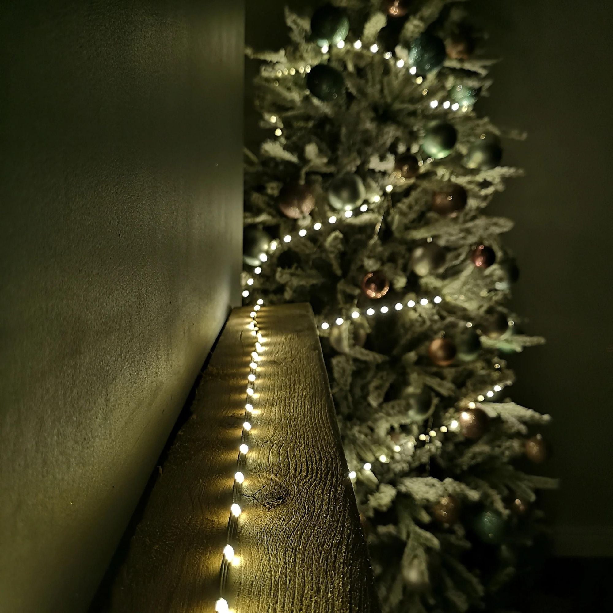 Warm white LED Christmas lights on a wooden surface, illuminating a frosted evergreen tree adorned with colorful ornaments.