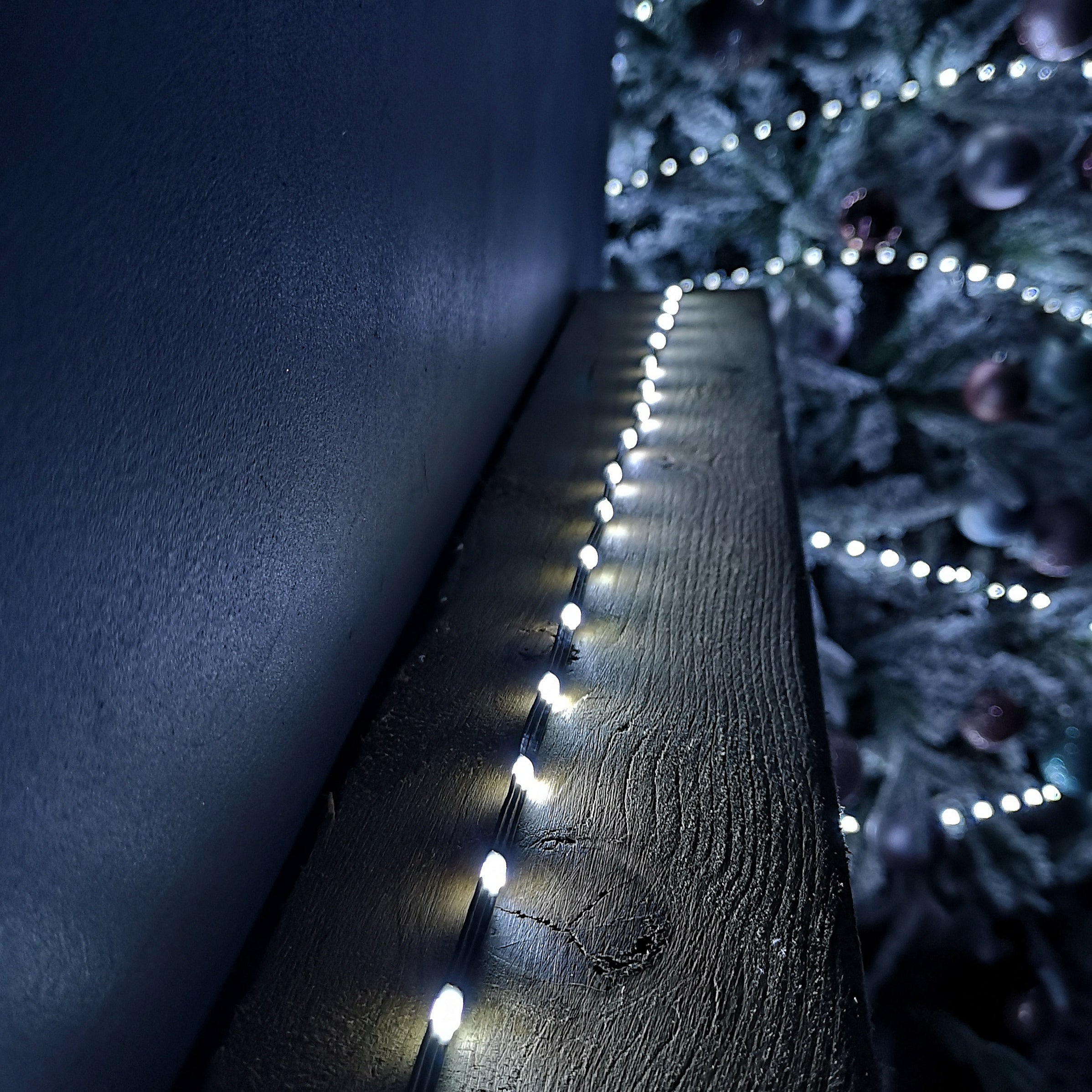 Flexible white Christmas lights with 500 LEDs, designed for indoor and outdoor use, displayed along a textured wooden surface against a backdrop of a decorated tree.