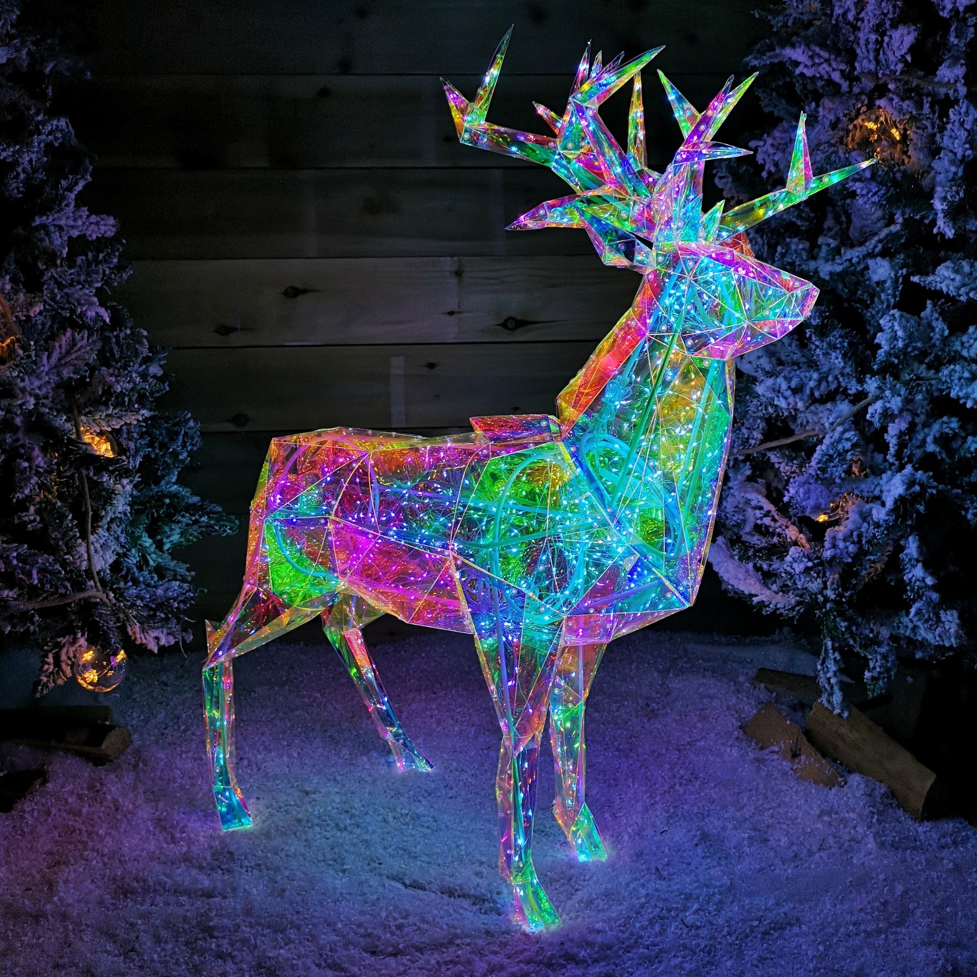 90cm Christmas stag made of translucent material with a geometric design, illuminated by 500 white LEDs, surrounded by snowy decor and evergreen trees.