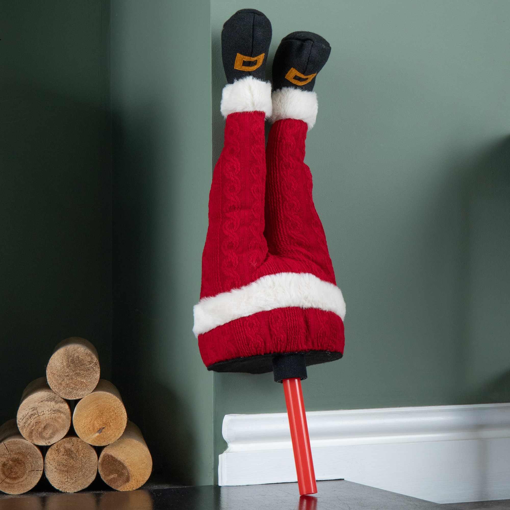 Soft animated Santa decoration with red knitted legs, black boots, and white fur trim, positioned upside down against a wall, with a bright orange pole.