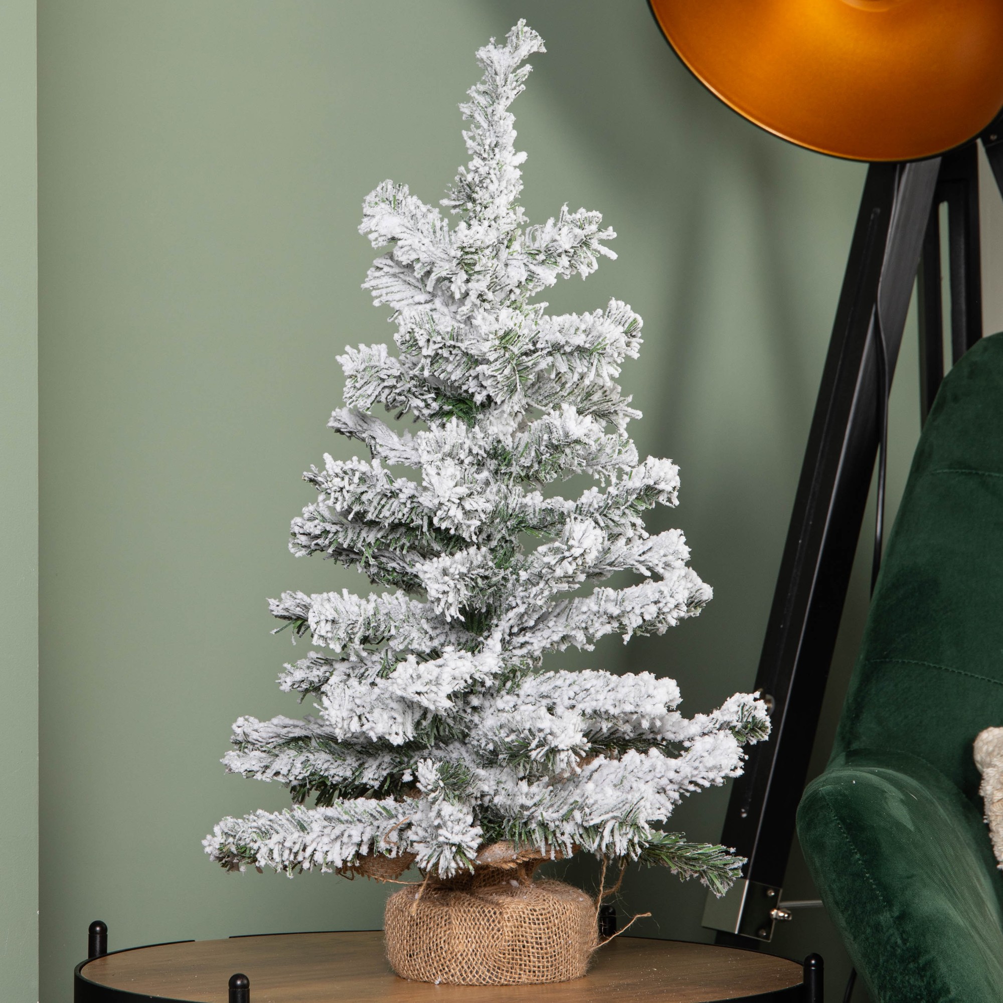 60cm mini Christmas tree with snow flocking, featuring green branches and a burlap base, displayed next to a modern lamp and a green armchair against a muted wall.