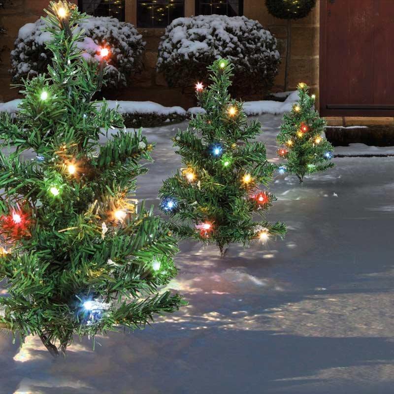 Six small artificial green Christmas trees with multi-colored LED lights, placed on a snowy outdoor path, creating a festive ambiance.