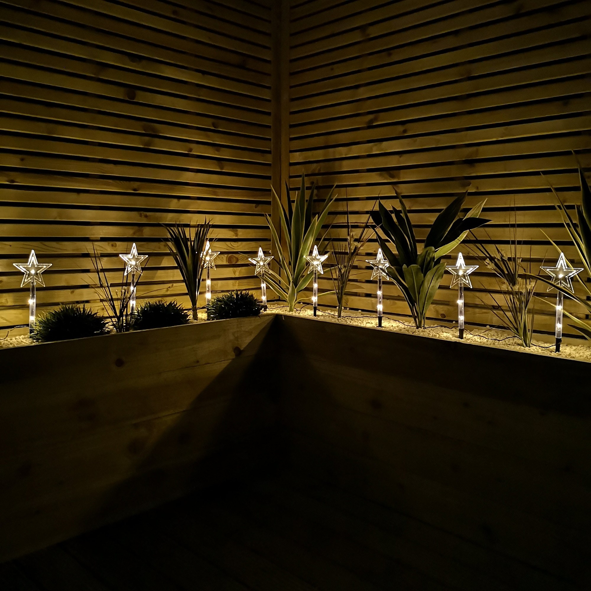 Set of 8 battery-operated LED multi-colored star path lights featuring clear star shapes on transparent stakes, illuminating a wooden backdrop with greenery.