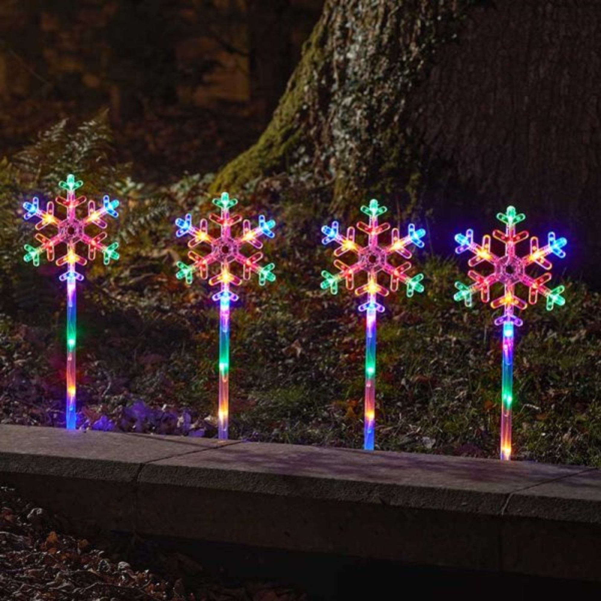 Four 43cm battery-powered snowflake path lights with multi-coloured LED lights, featuring intricate snowflake designs and clear plastic stakes, set against a natural backdrop.