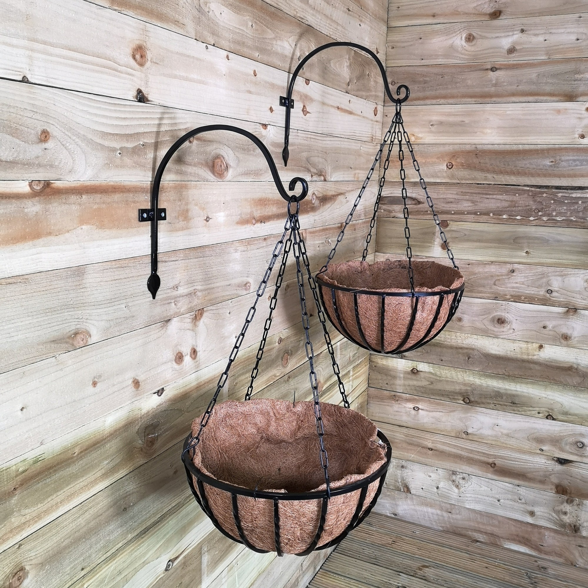 Two black metal hanging baskets with a twisted bar design, featuring coco fibre liners, suspended from hooks against a wooden wall. Each basket measures 40cm in diameter.