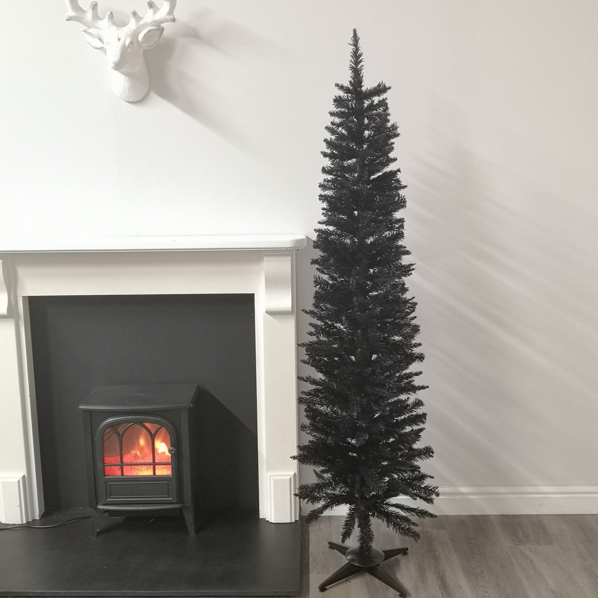 6ft black pencil pine artificial Christmas tree with 321 tips, standing beside a black electric fireplace, set against a light-colored wall.