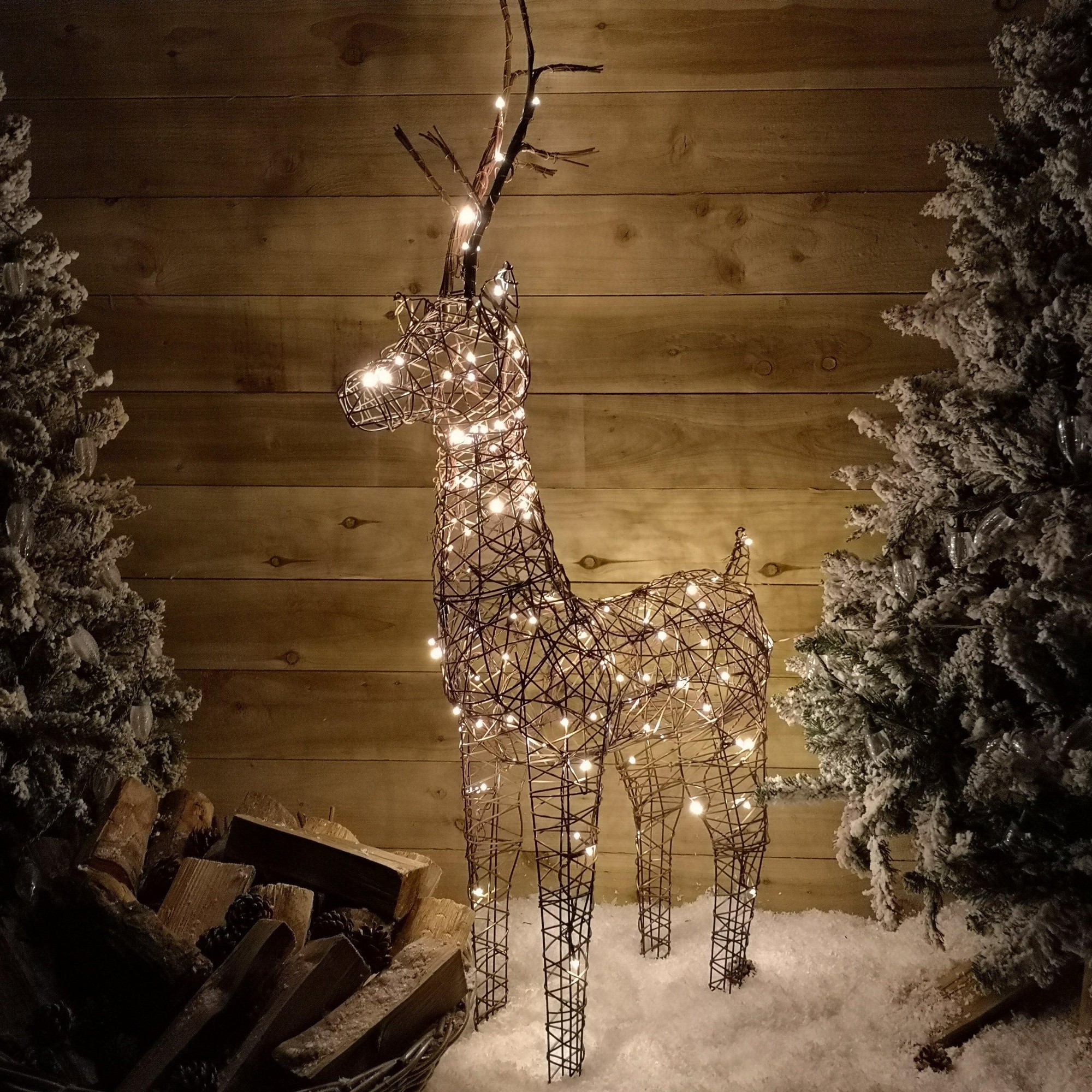 Brown wicker reindeer decoration standing 53 inches tall, adorned with warm LED lights, set against a snowy backdrop with evergreen trees and wooden wall.