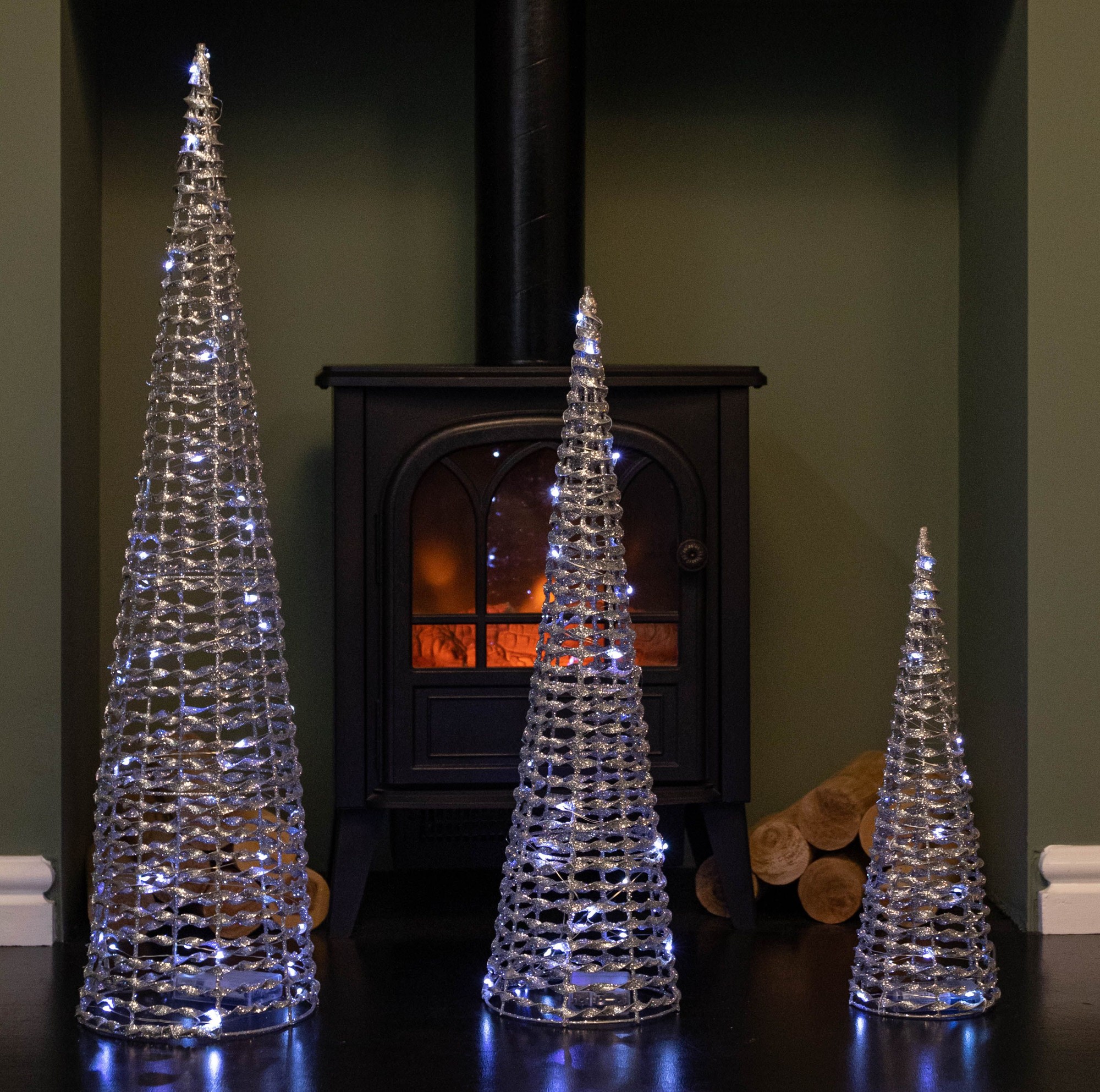 Set of 3 silver glitter cone trees featuring a twisted design, adorned with 90 cool white LEDs, displayed beside a black wood stove against a green wall.