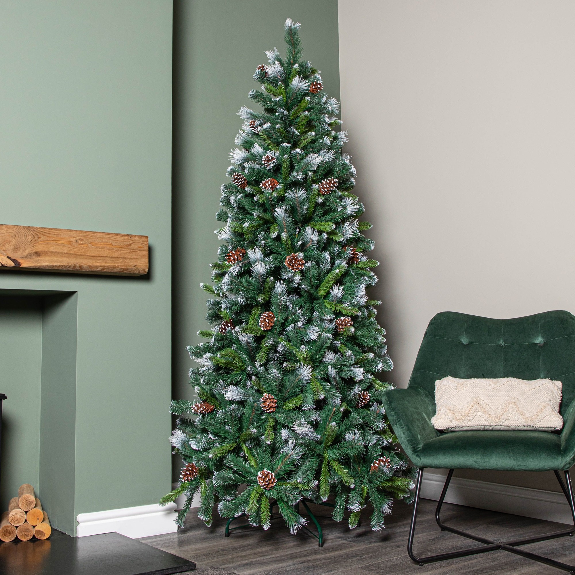 7ft frosted glacier pine Christmas tree with snow-tipped branches, pinecones, and 956 tips, set against a green wall and accompanied by a cozy chair and fireplace.