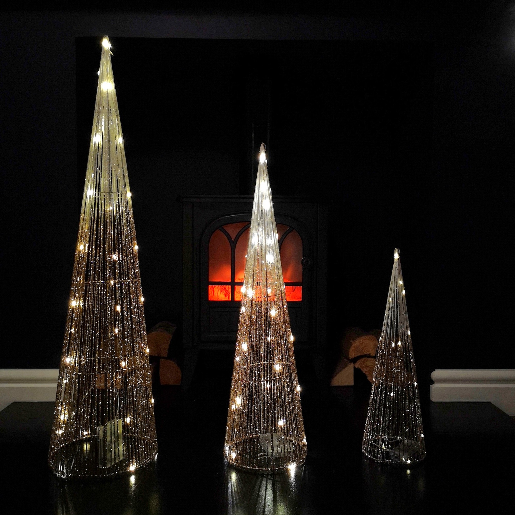 Three gold LED tree cone obelisks of varying heights, adorned with 90 warm white lights, set against a dark background with a glowing fireplace.