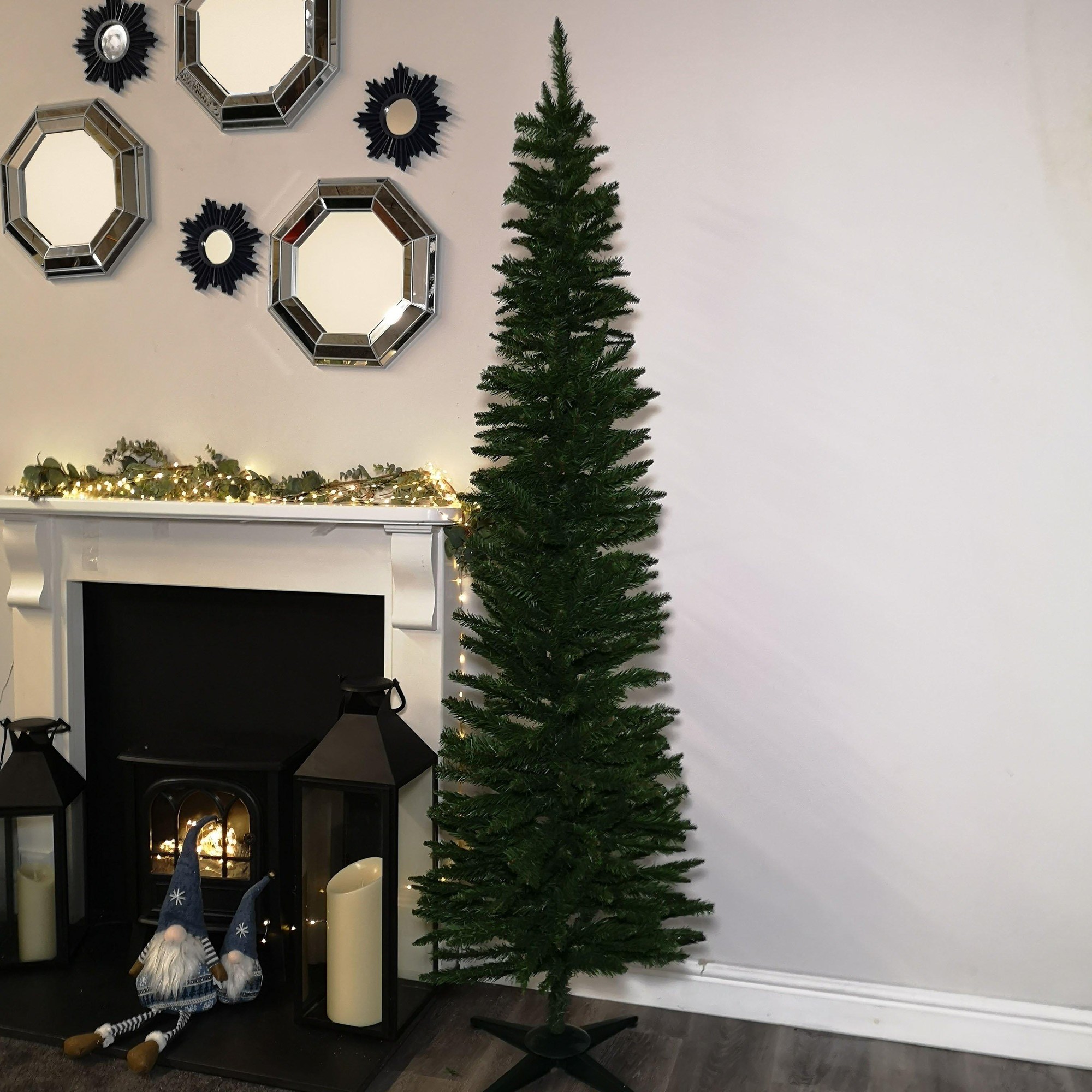 7ft green pencil style slim artificial Christmas tree with dense branches, set against a light wall near a fireplace, complemented by decorative mirrors and festive lights.