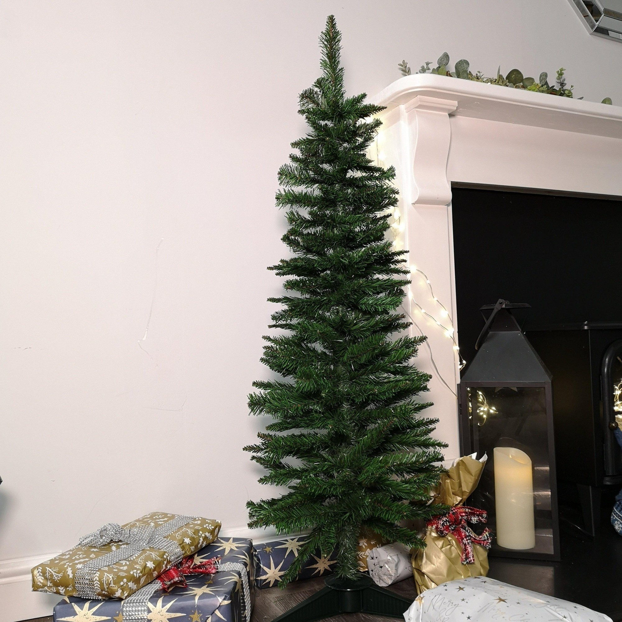 4ft slim artificial Christmas tree in green with a pencil style design, placed beside wrapped gifts and a lantern, featuring a realistic foliage appearance.