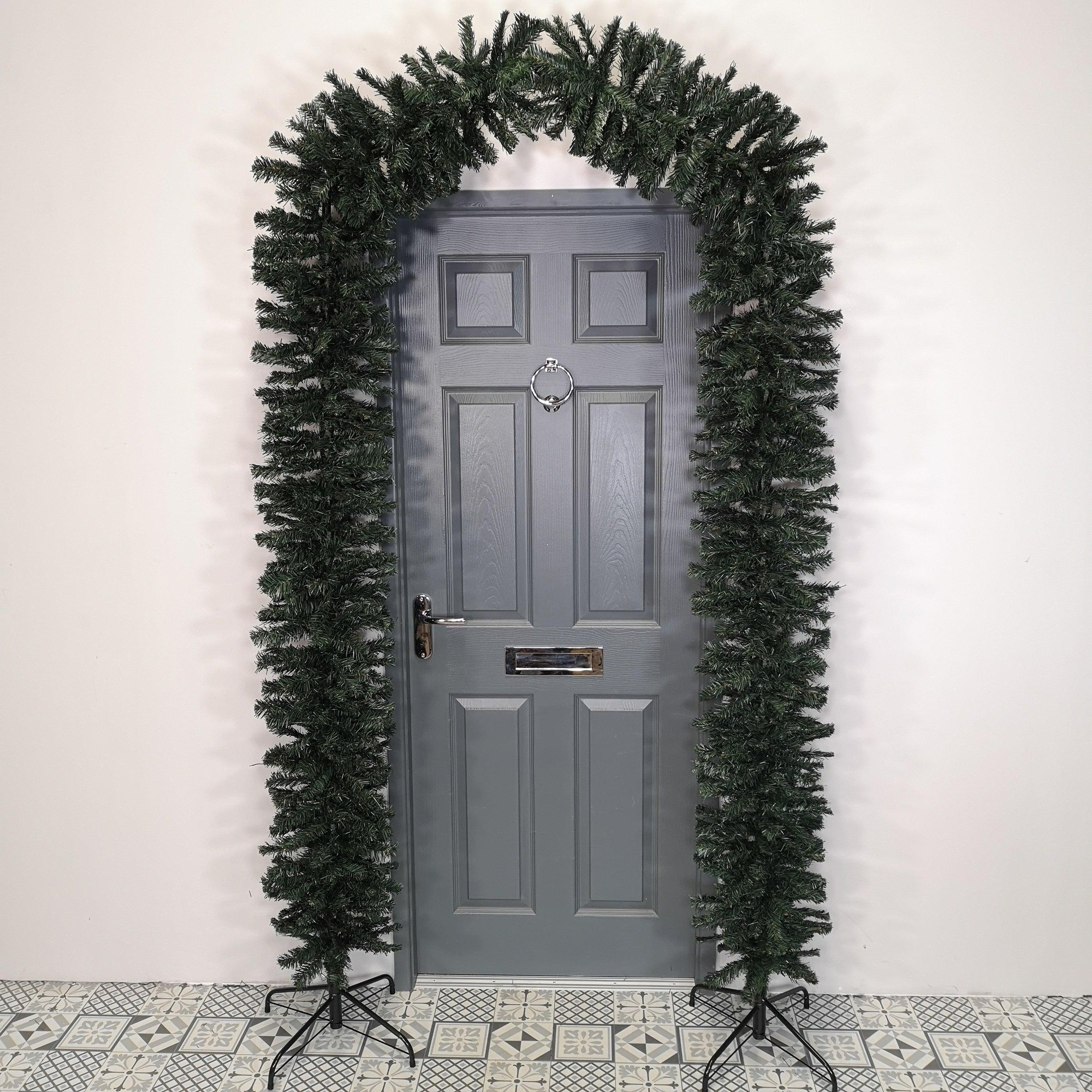 Tall 2.4M green artificial Christmas tree arch with a single door frame design, featuring lush foliage and sturdy stands, set against a gray door and patterned floor.