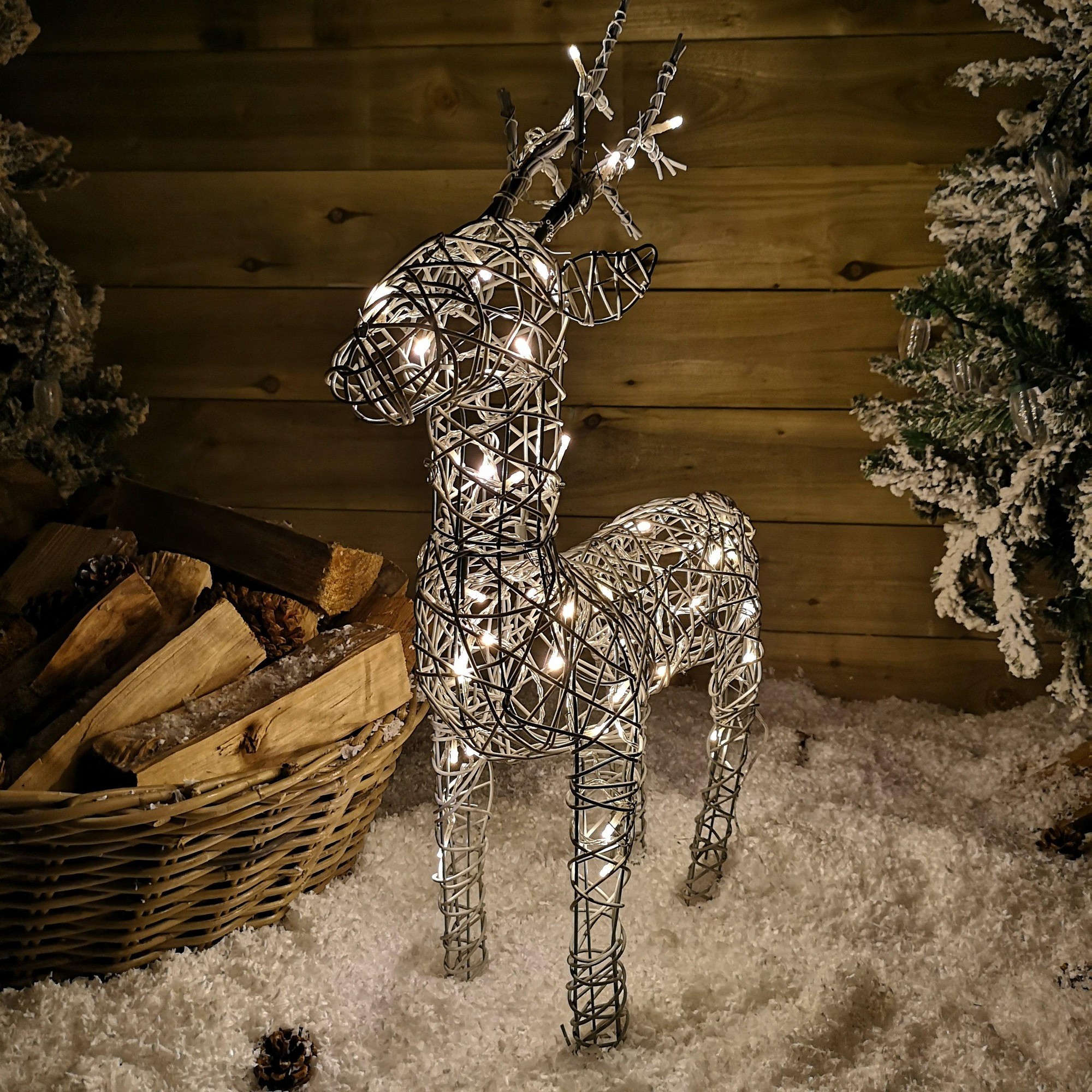 60cm grey wicker reindeer decoration with LED lights, featuring a detailed woven design and antlers, set against a snowy backdrop with wooden logs and pine trees.