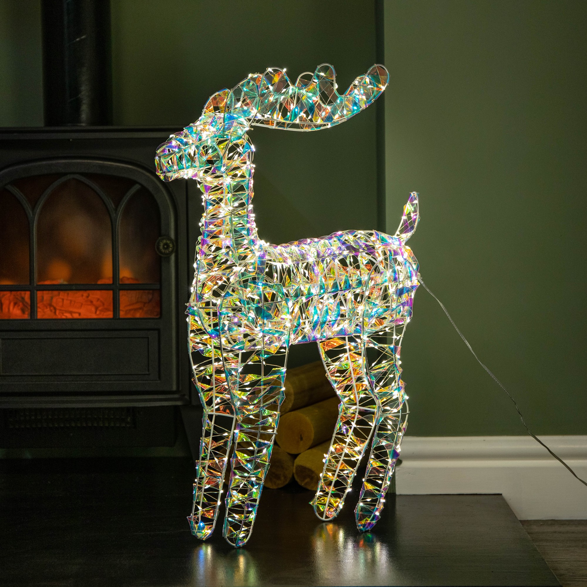 60cm iridescent shimmer Christmas reindeer made of wire, adorned with 720 warm white LEDs, standing beside a fireplace against a green wall.