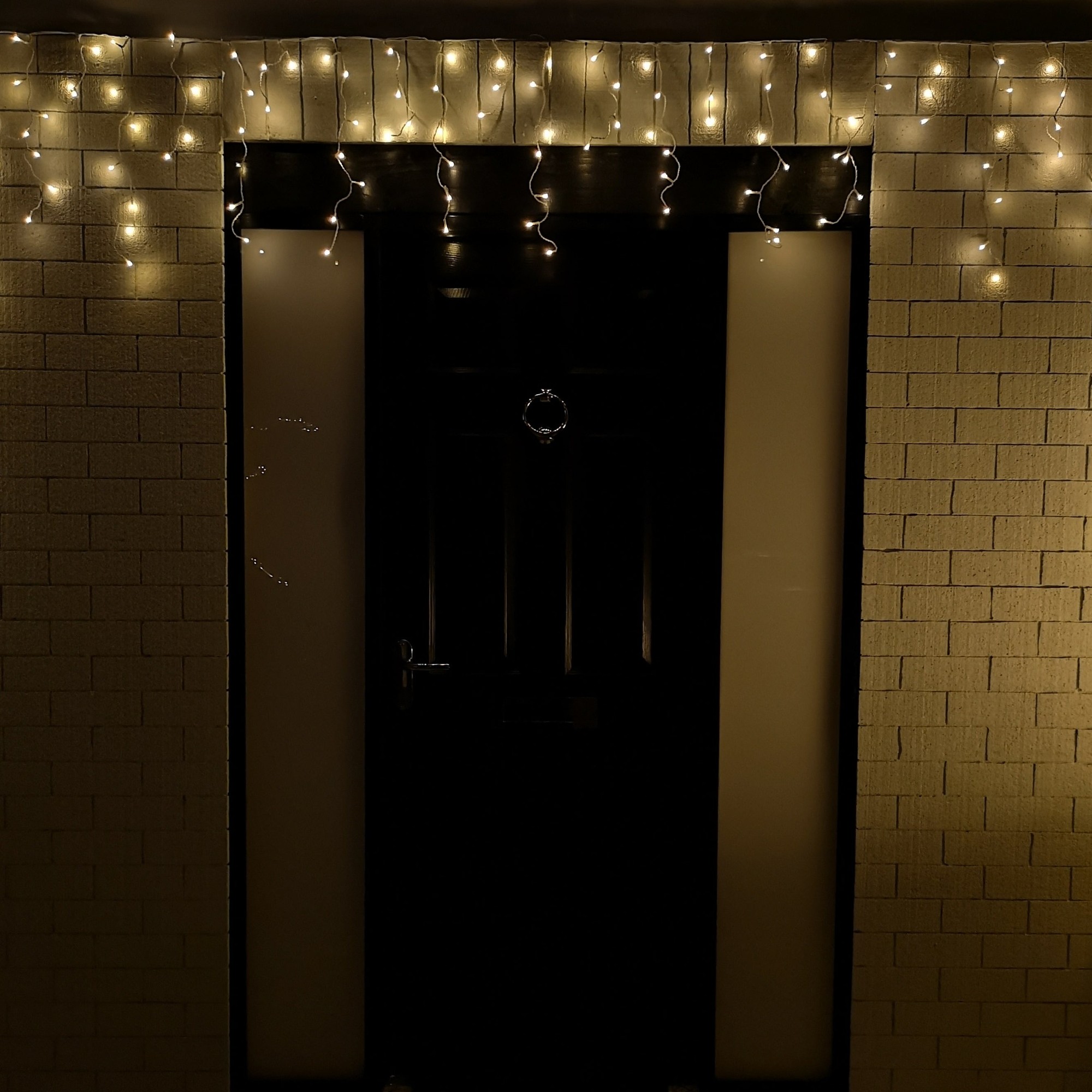 Warm white LED frosted icicle lights hang above a dark door, creating a festive ambiance with a timer feature, illuminating the textured wall in a cozy glow.