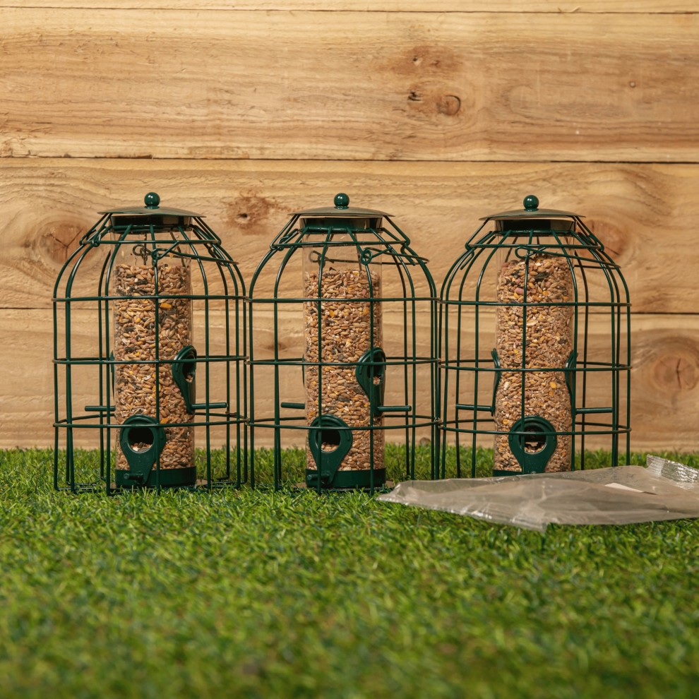 Three green metal hanging bird nut feeders with a squirrel guard design, featuring cylindrical seed holders and a pack of nut feed on grass against a wooden background.