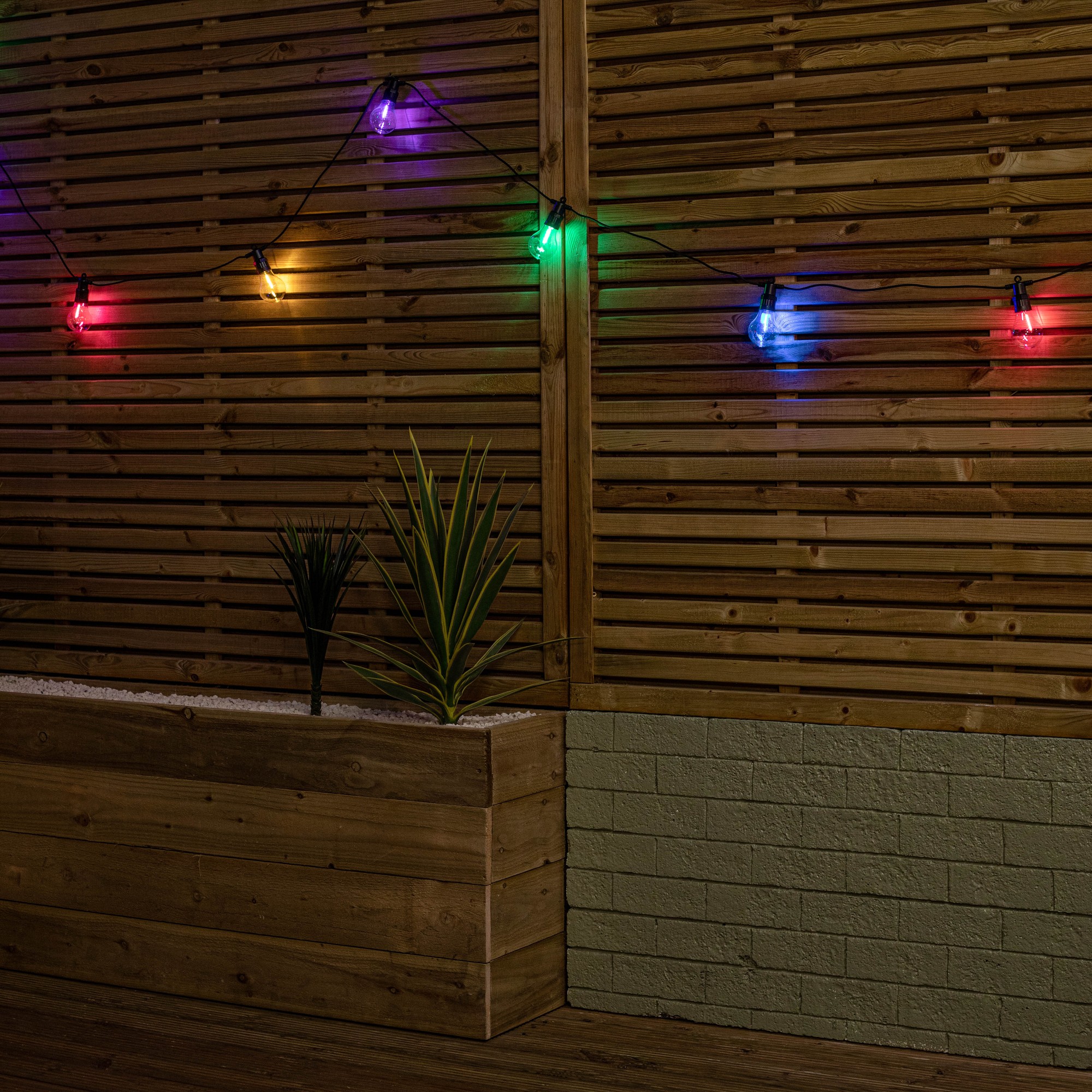 Connectable festoon lights featuring multi-coloured LEDs in vibrant hues, hanging against a wooden backdrop, with a planter and greenery below.
