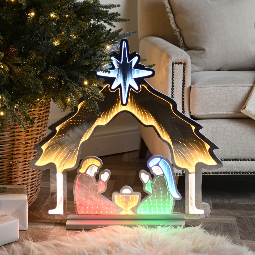 65cm light-up nativity scene featuring a stable with a blue star, illuminated figures of Mary, Joseph, and baby Jesus, enhanced by multi-coloured LEDs, set near a Christmas tree.