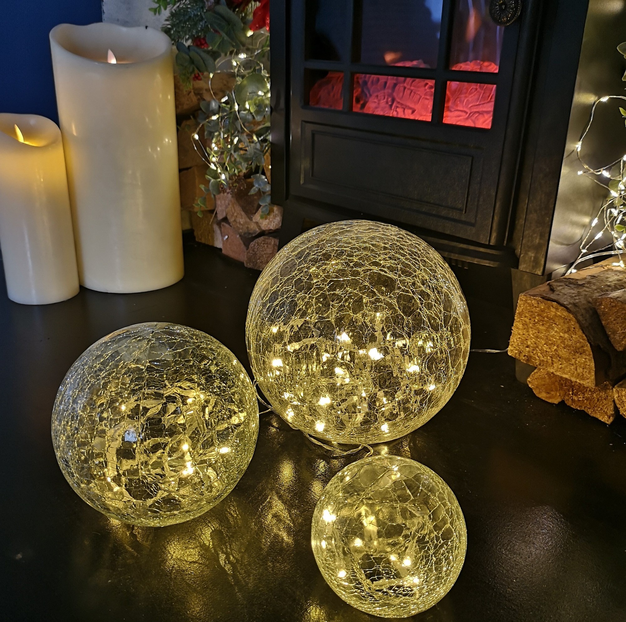 Set of three oval glass balls in warm white, featuring a crackled texture and 60 LED lights, arranged beside candles and a fireplace for festive decoration.